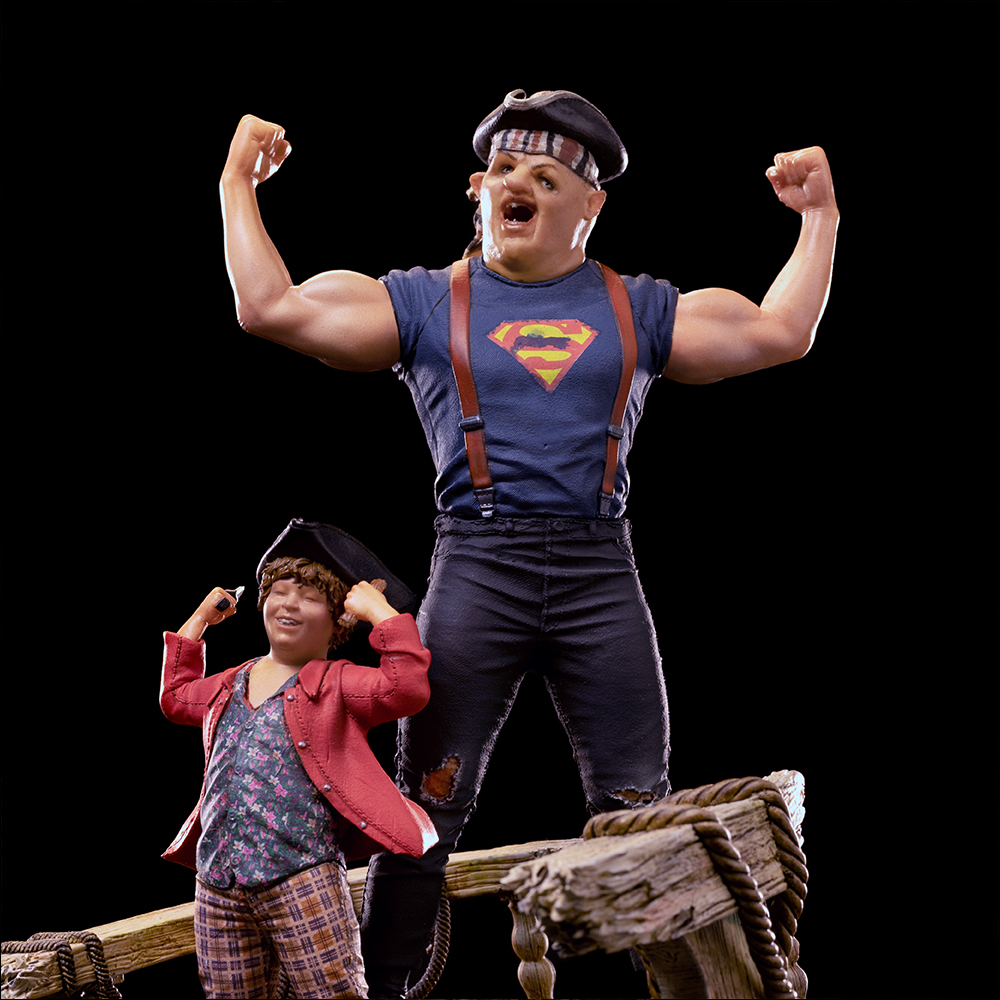 Zwei Figuren stehen auf Holz. Eine muskulöse Figur in einem Superman-Shirt, Arme erhoben. Die andere trägt eine rote Jacke und Hut, lächelnd.