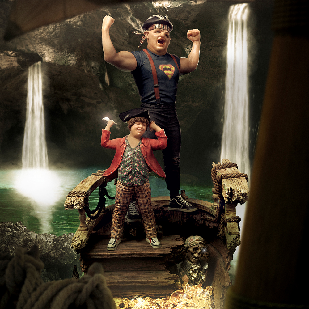 Zwei Figuren auf einem Schiff, Wasserfälle im Hintergrund. Eine Figur ist muskulös mit Superman-Shirt, die andere in einem roten Mantel.