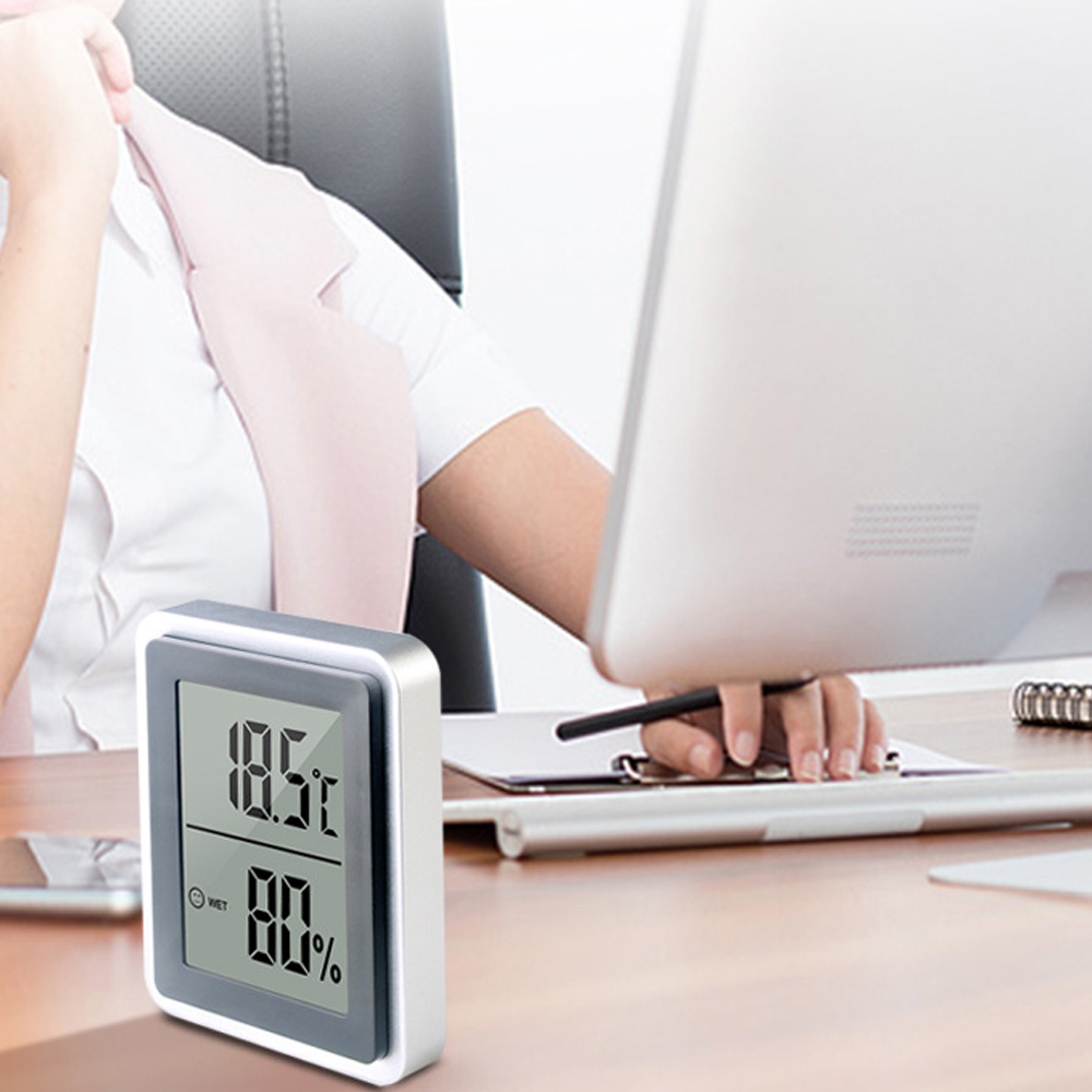 Eine Schreibtischszene mit einem digitalen Thermometer, das 18,5 °C und 80 % Luftfeuchtigkeit anzeigt. Im Hintergrund arbeitet eine Person am Computer.