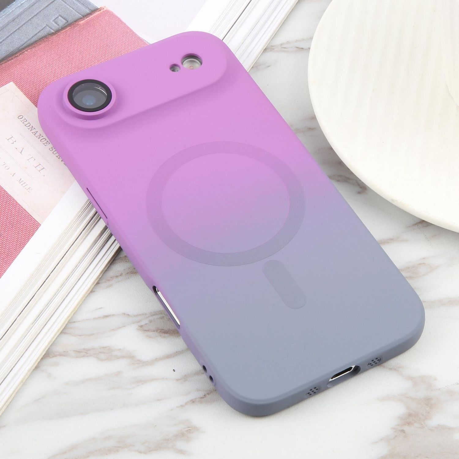 Una funda de teléfono con degradado púrpura-gris sobre una superficie de mármol. Cerca, un libro y un plato blanco.