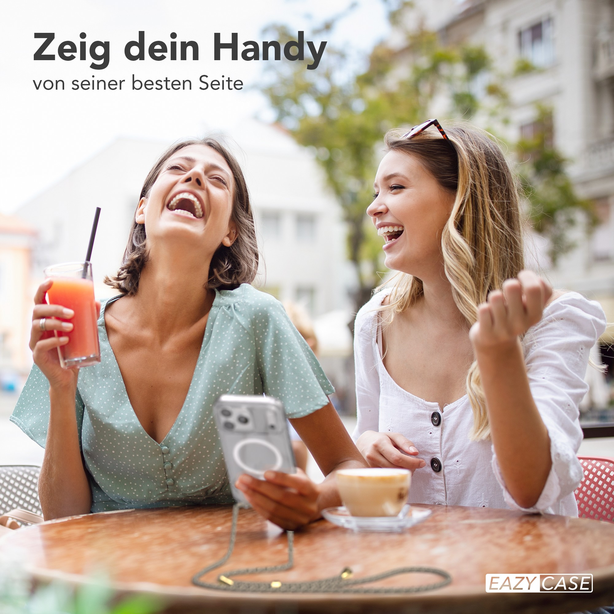 Zwei lachende Frauen in einem Straßencafé. Eine hält ein Handy mit Band. Getränke und Kaffee auf dem Tisch.