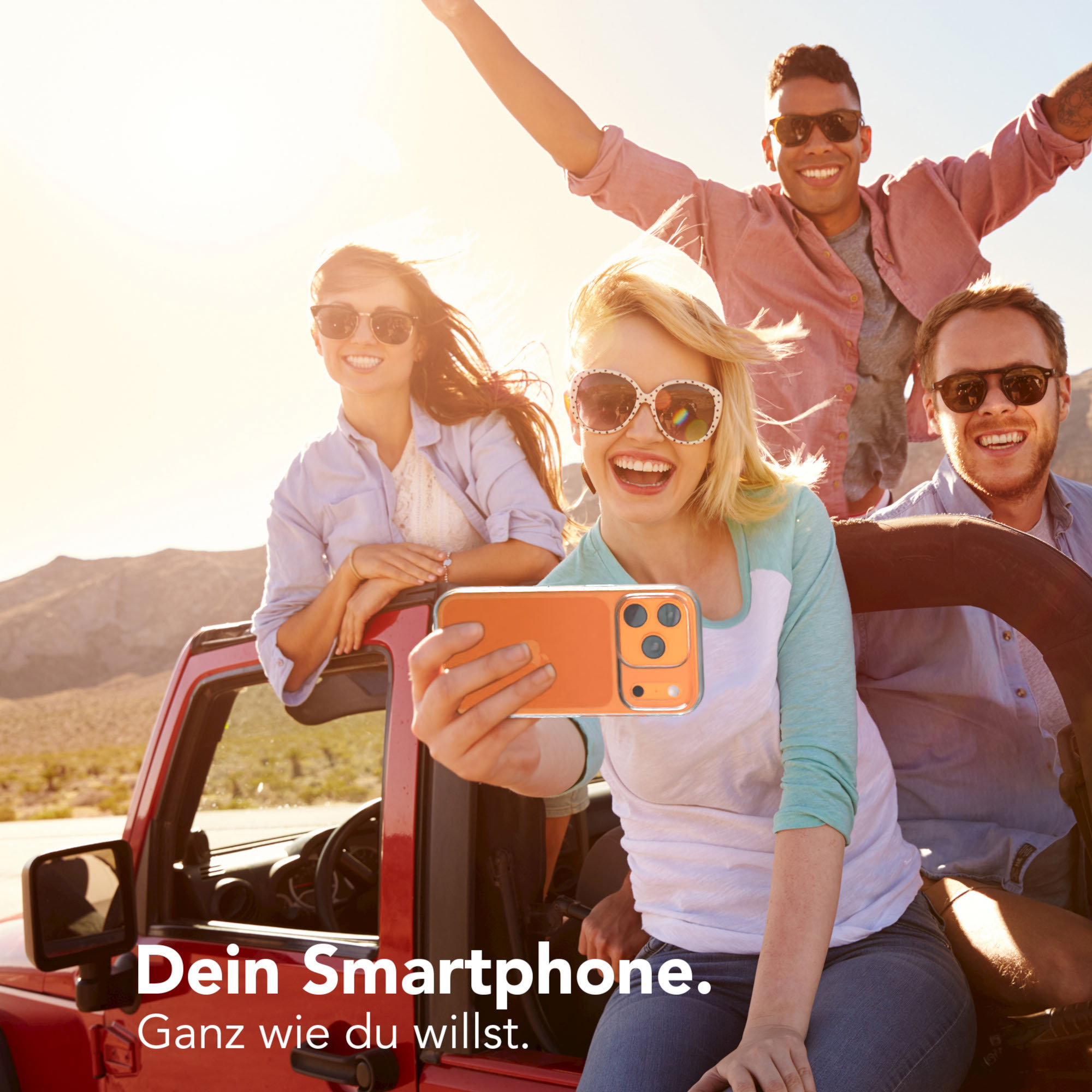 Vier Personen in einem roten Jeep machen ein Selfie. Sie tragen Sonnenbrillen und lächeln. Der Hintergrund ist eine Wüste.