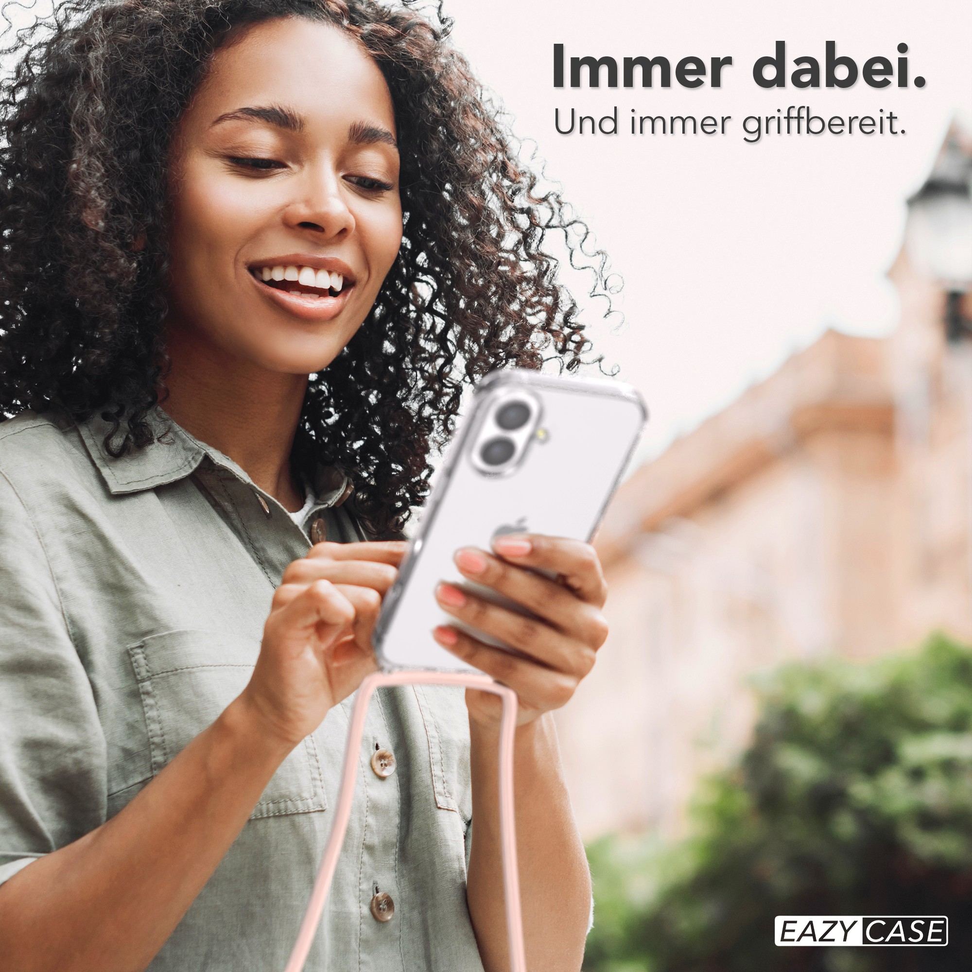 Lächelnde Frau mit Handy in transparenter Hülle. Rosa Träger und verschwommener Hintergrund mit Gebäude und Grün.
