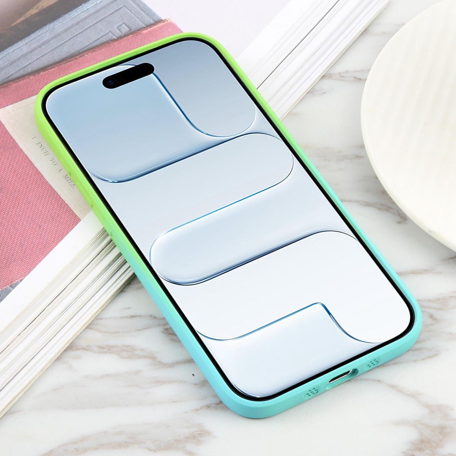 Ein Smartphone in einer grün-blauen Hülle liegt auf einer Marmoroberfläche, neben einem Buch und einem weißen Teller.