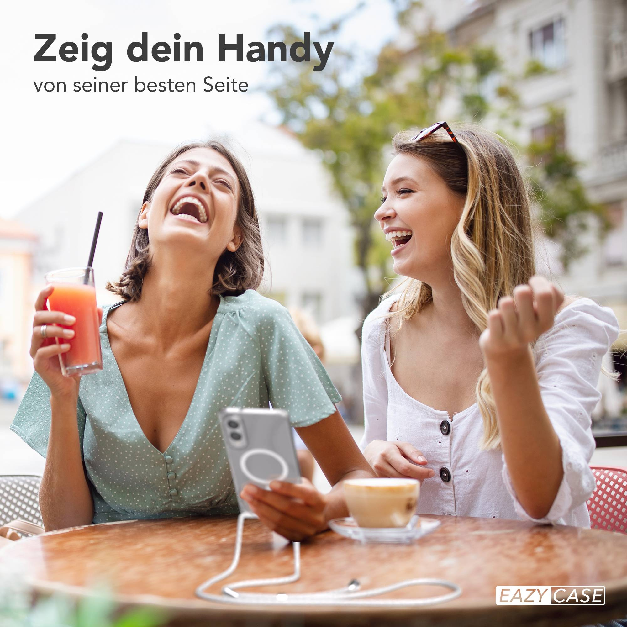 Zwei lachende Frauen an einem Tisch. Eine hält ein Telefon, die andere ein Getränk. Café, sonniger Tag.