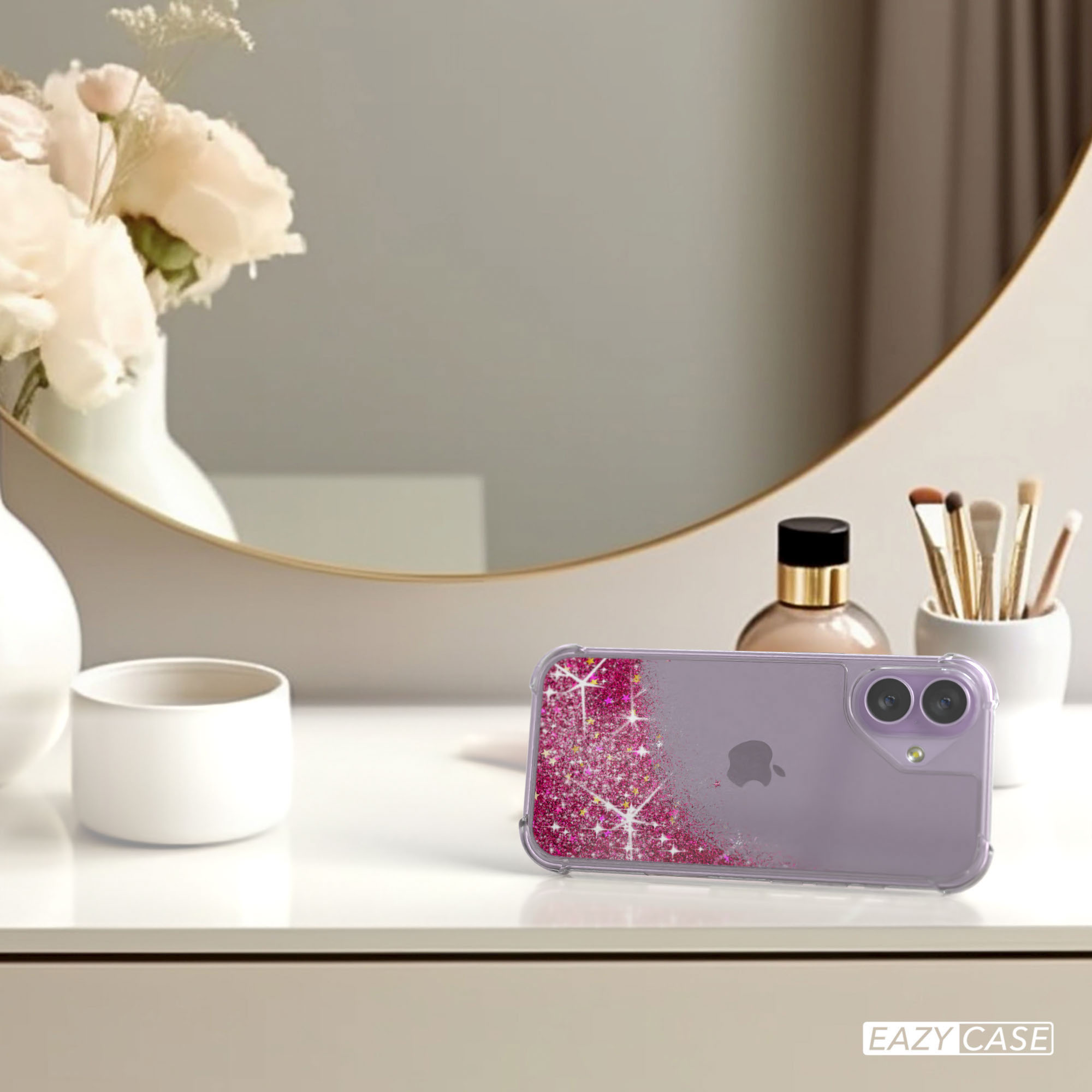 Ein iPhone in einer rosa Glitzerhülle liegt auf einem weißen Tisch. Blumen und ein Spiegel sind im Hintergrund. Make-up-Pinsel sind sichtbar.