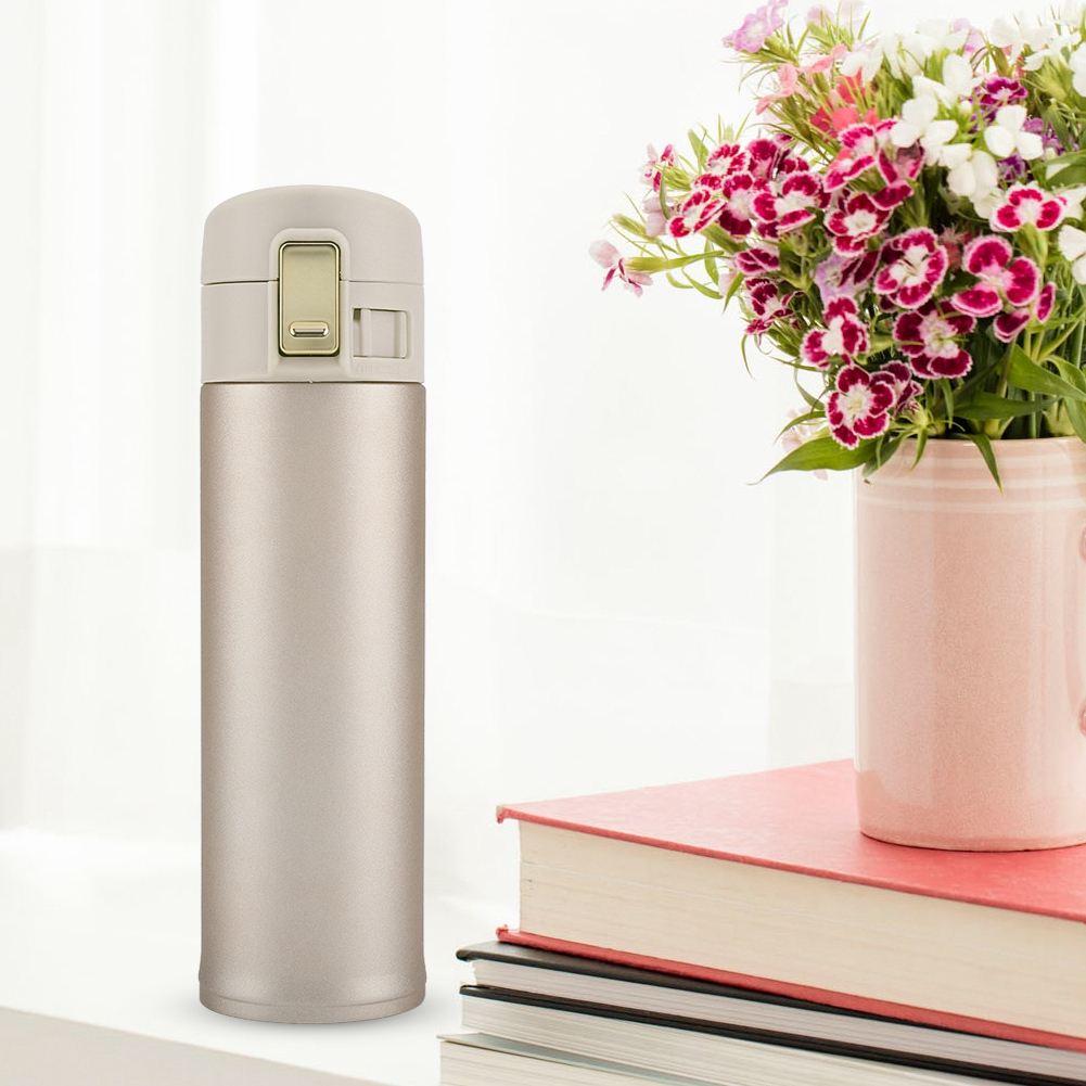 Eine metallische Thermosflasche steht neben einer Vase mit Blumen auf einem Stapel Bücher. Der Hintergrund ist weiß.