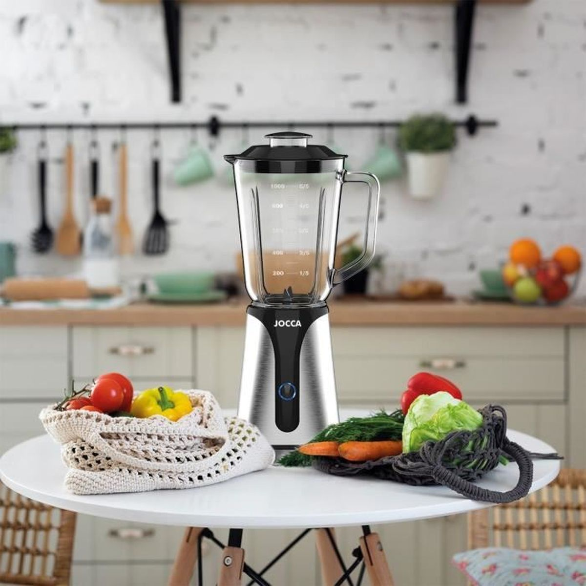 Mixer auf einem weißen Tisch mit Gemüse in Netztaschen. Hintergrund zeigt Küchenutensilien und ein Regal mit Pflanzen.