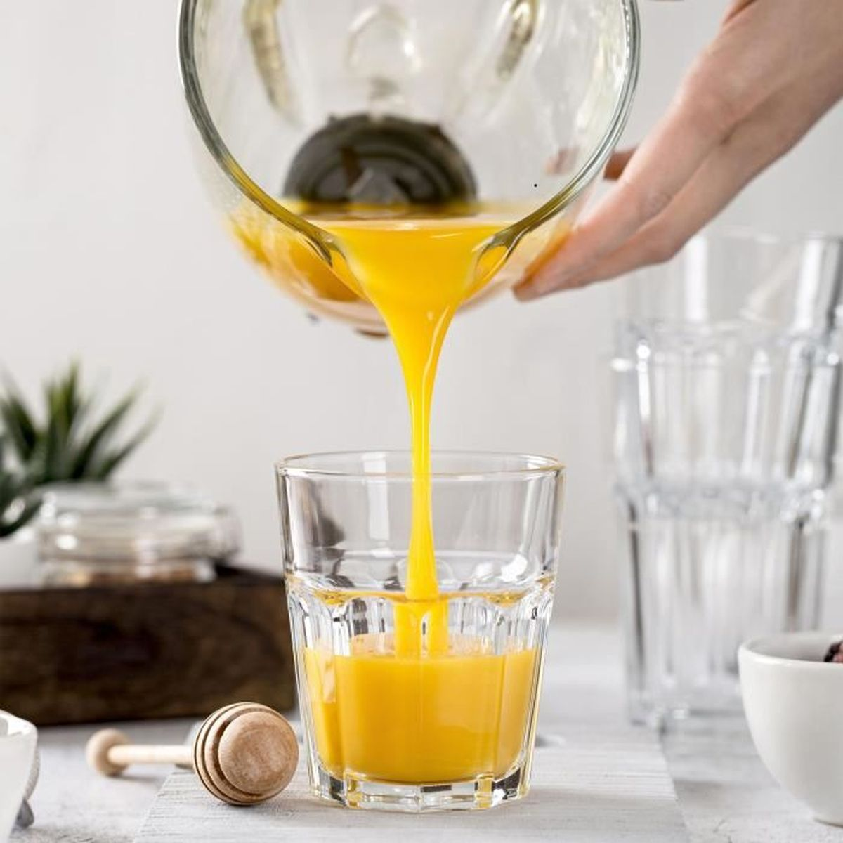 Gelbe Flüssigkeit gießt aus einem Krug in ein Glas. Honiglöffel und andere Gläser sind im Bild.