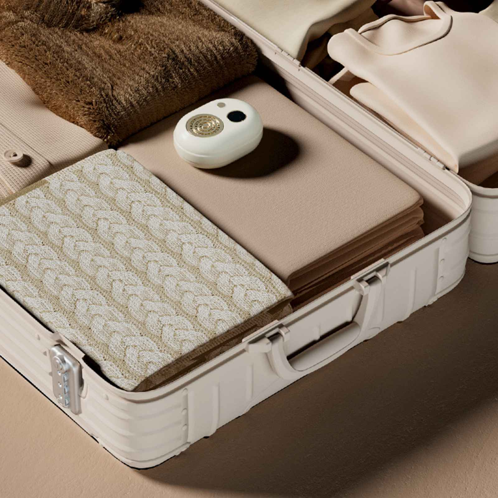 Offener Koffer gefüllt mit beigefarbenen Gegenständen: gefaltete Kleidung, eine Zopfstrickdecke und ein weißes elektronisches Gerät.