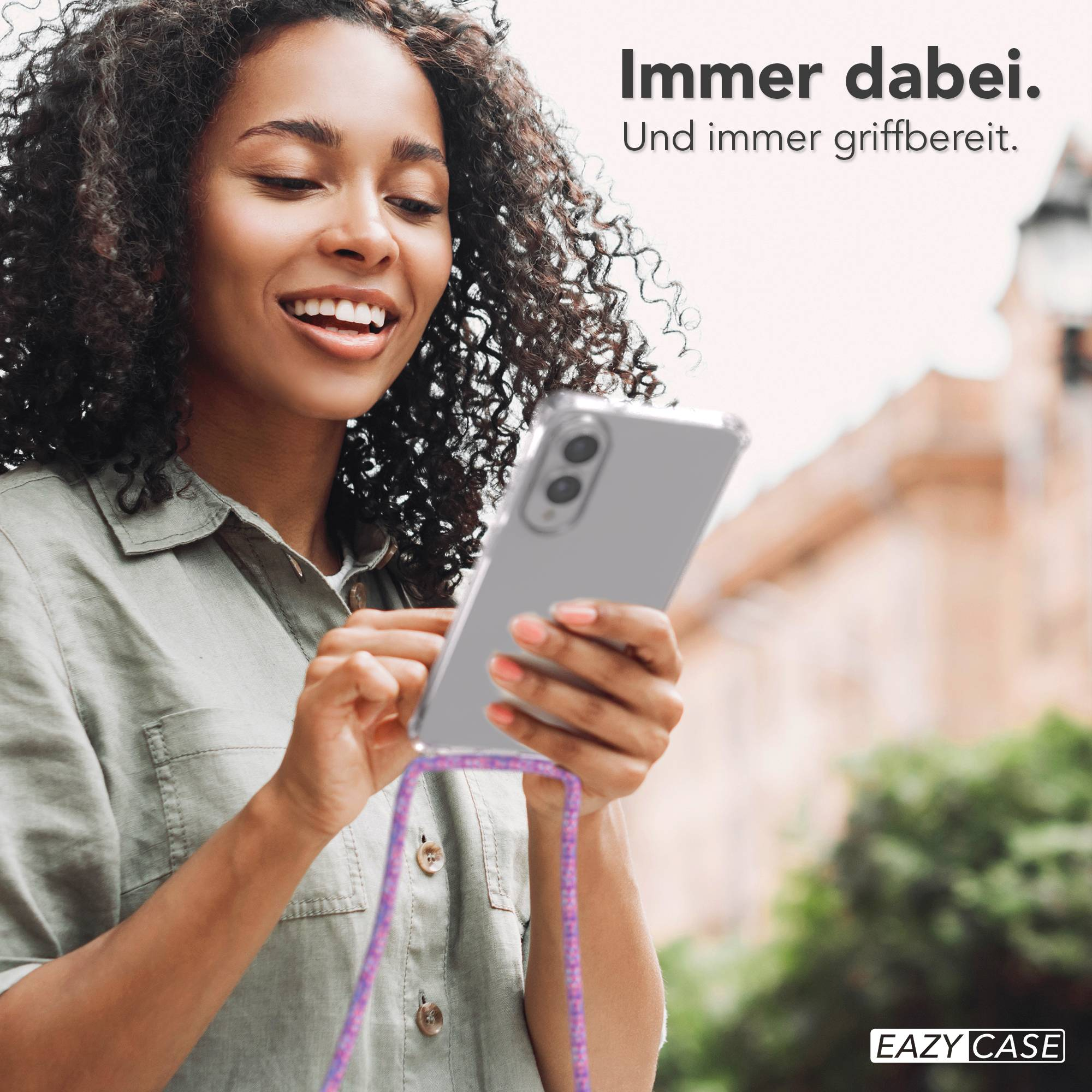 Eine Frau hält ein Telefon mit Gurt. Sie lächelt, mit verschwommenen Gebäuden im Hintergrund.