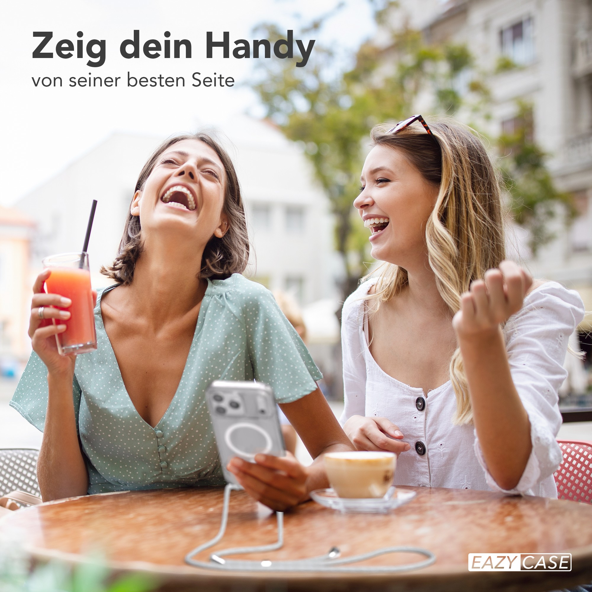 Zwei lachende Frauen in einem Café, eine hält ein Telefon mit Gurt, die andere ein Getränk. Hintergrund Gebäude.
