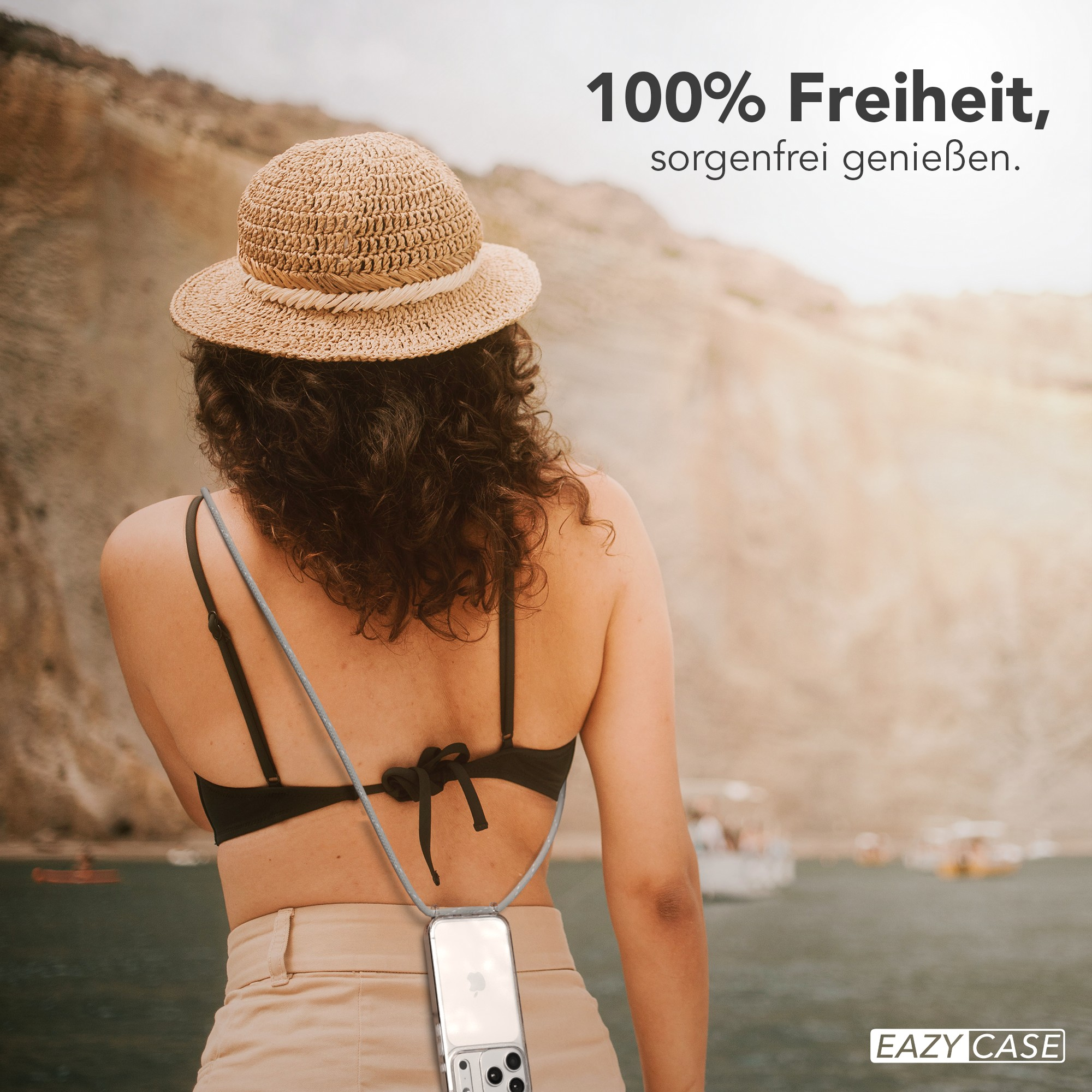 Frau mit Hut, schwarzem Oberteil und beiger Hose. Eine Handyhülle hängt um ihren Hals, landschaftlicher Hintergrund.