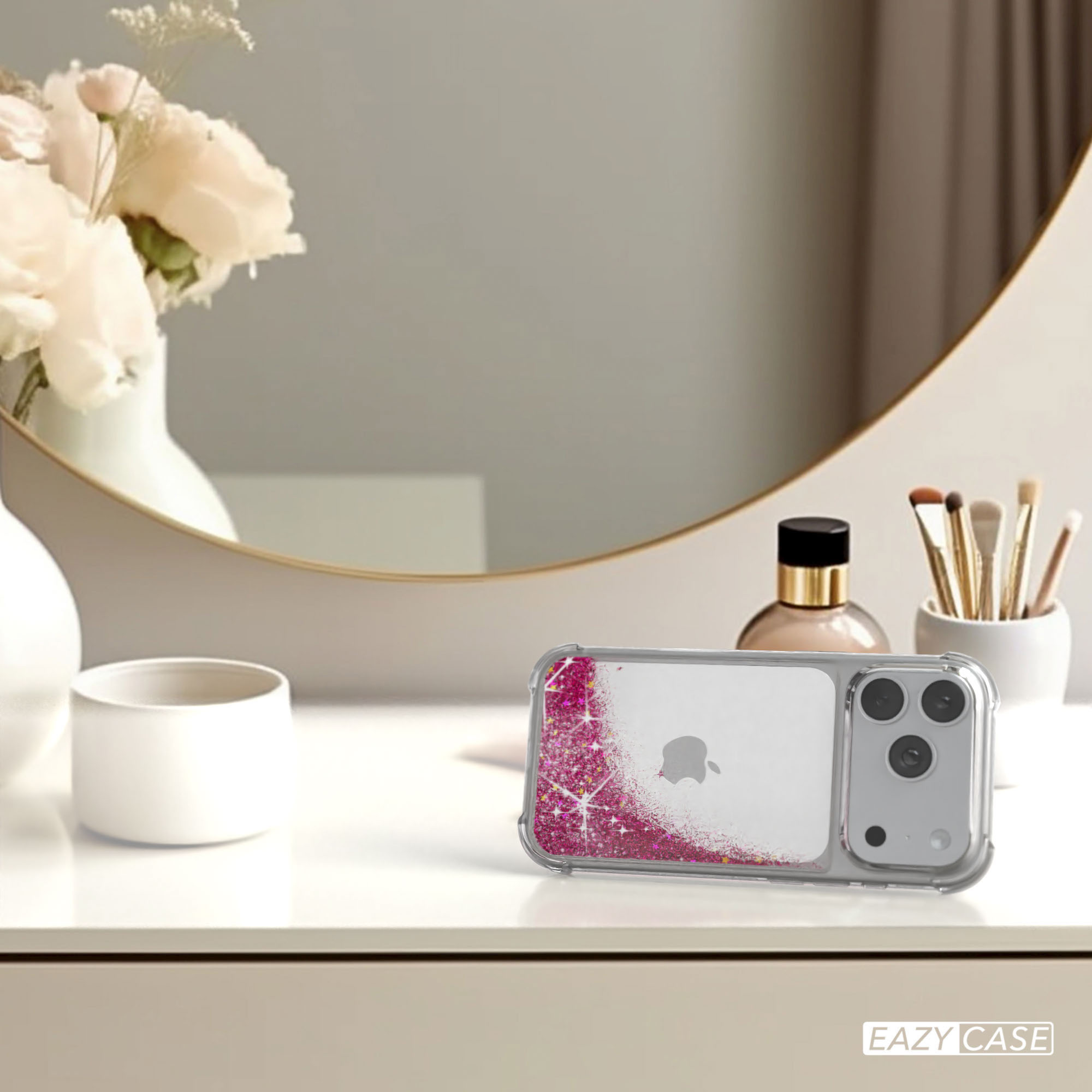 Ein iPhone in einer Hülle mit pinkfarbenem Glitzer liegt auf einer weißen Oberfläche. Blumen, ein Spiegel und Make-up sind im Hintergrund.