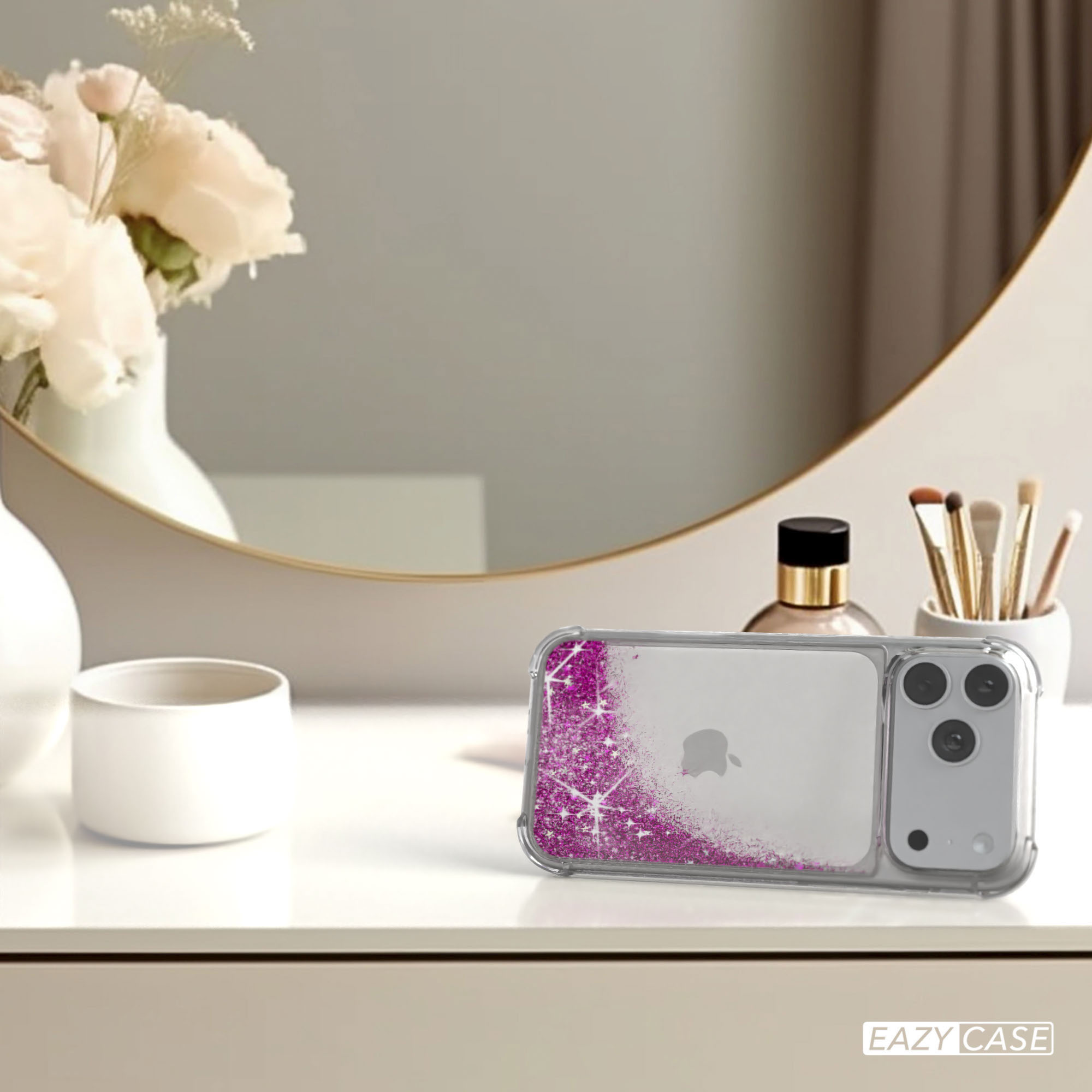 Ein iPhone in einer pinkfarbenen Glitzerhülle liegt auf einer weißen Oberfläche mit Spiegel, Blumen und Kosmetika im Hintergrund.