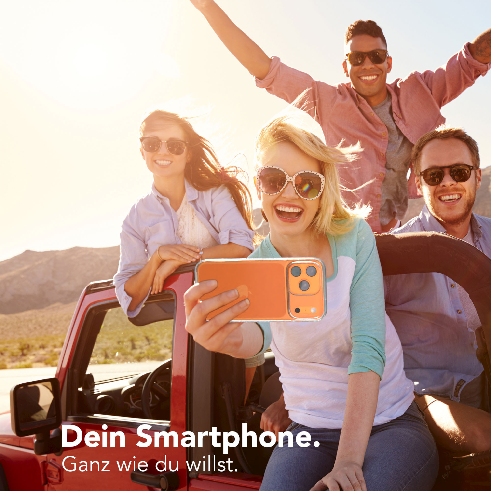 Vier Personen in einem roten Jeep machen ein Selfie mit einem orangenen Telefon. Sie lächeln. Berge sind im Hintergrund sichtbar.
