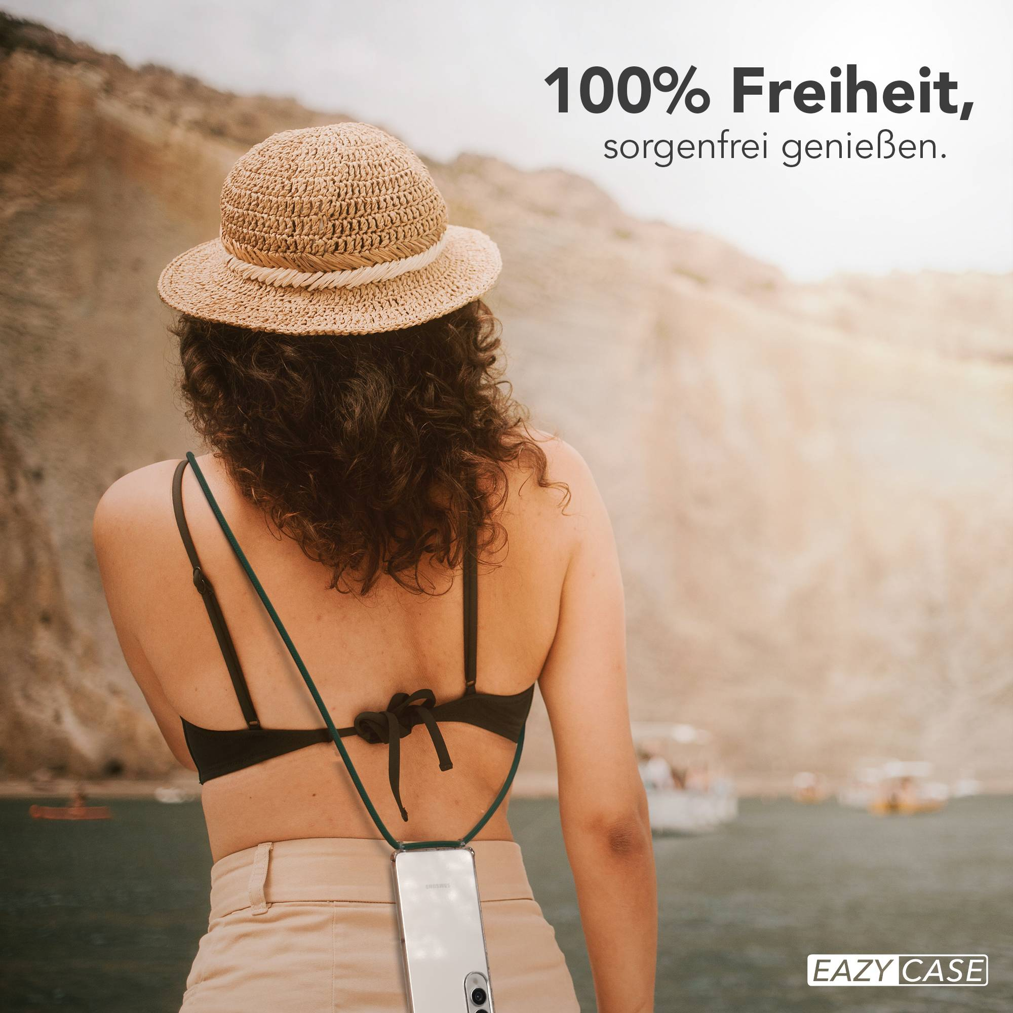 Frau mit Hut, schwarzem Oberteil und khakifarbener Hose, hält Handy mit grünem Riemen. Hintergrund: Meer und Berge.