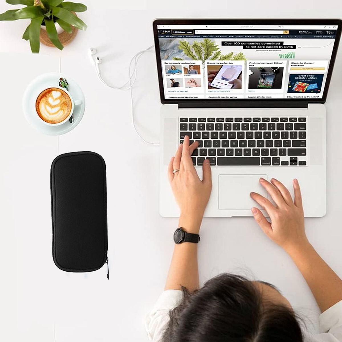 Eine Person benutzt einen Laptop. Es gibt eine Tasse Kaffee, eine schwarze Hülle und Kopfhörer. Der Laptop zeigt eine Amazon-Webseite.