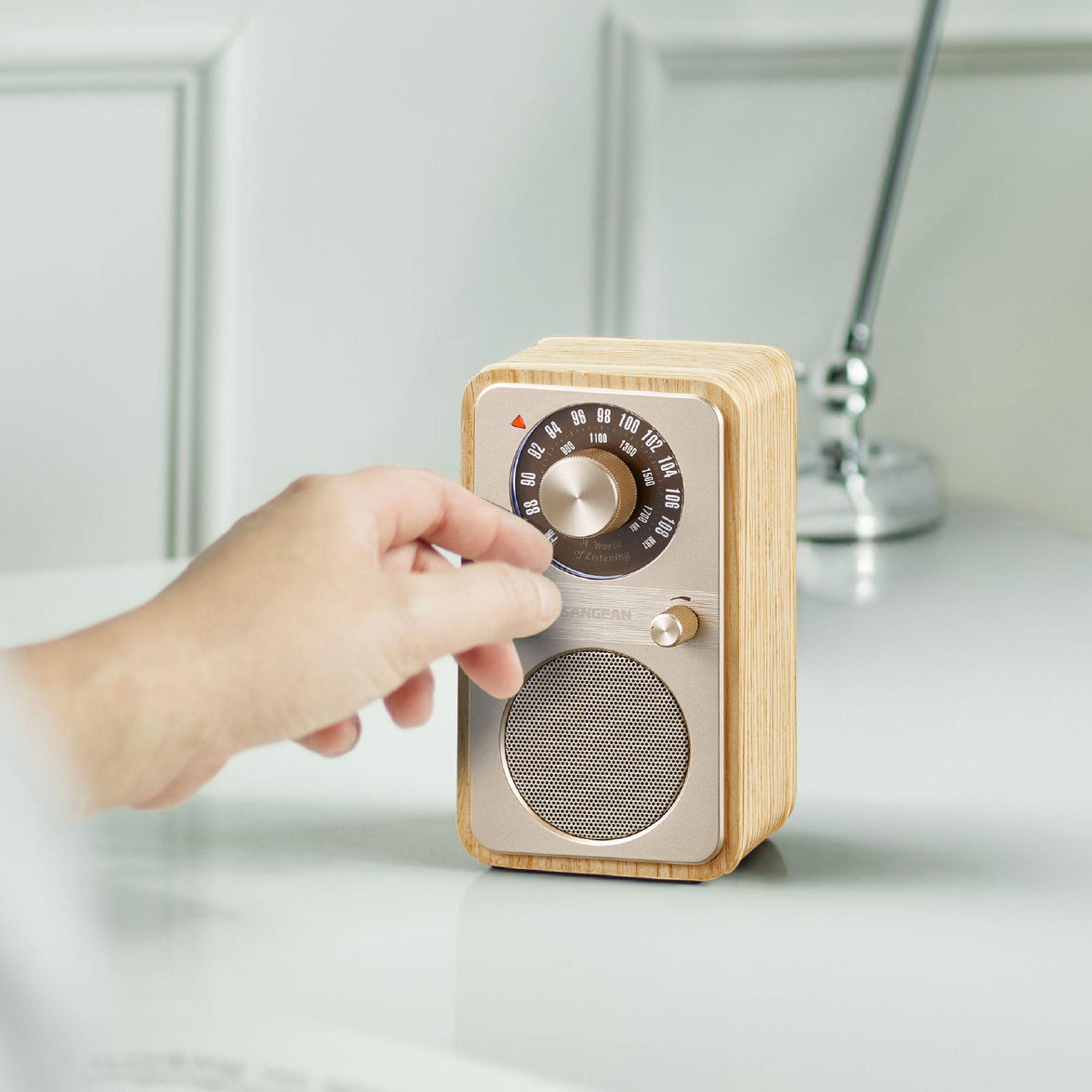 Eine Hand stellt den Drehregler eines Holzradios auf einer weißen Oberfläche ein. Das Radio hat einen Lautsprecher und ein Zifferblatt.