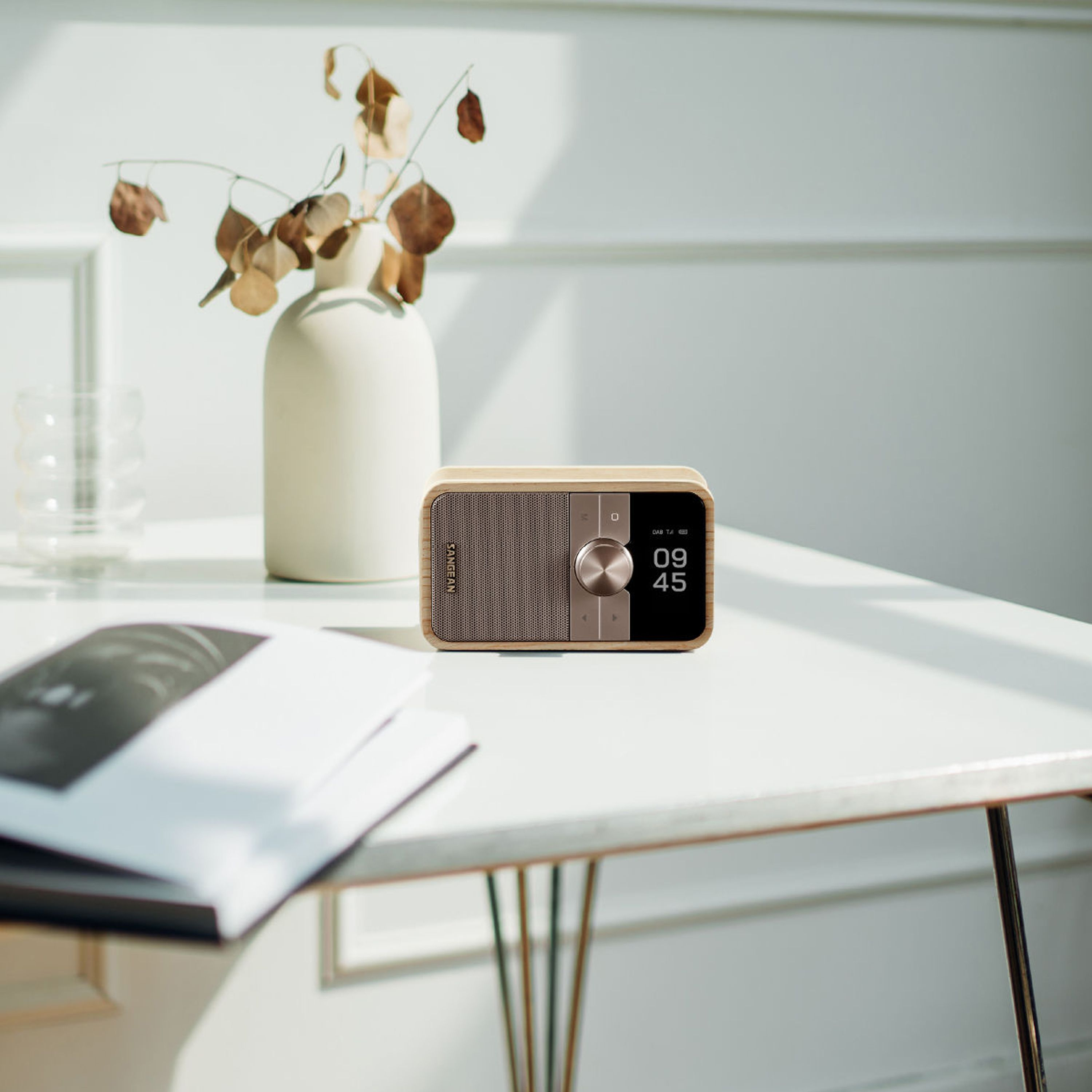 Una radio beige con display digitale (09:45) su un tavolo bianco. Dietro, un vaso con foglie secche.