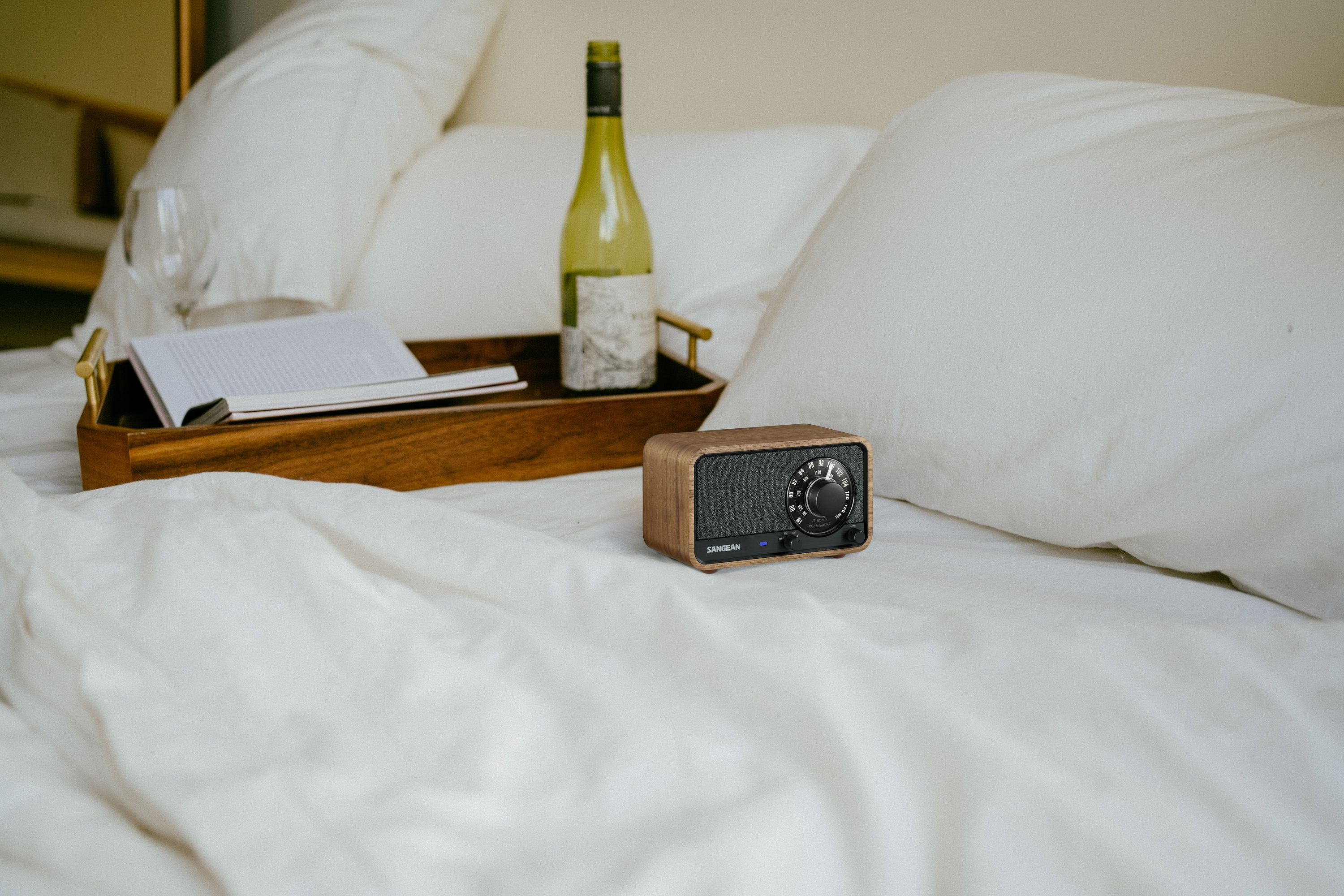 Ein Holzradio liegt auf einem weißen Bett, im Hintergrund eine Weinflasche und ein Notizbuch auf einem Tablett.
