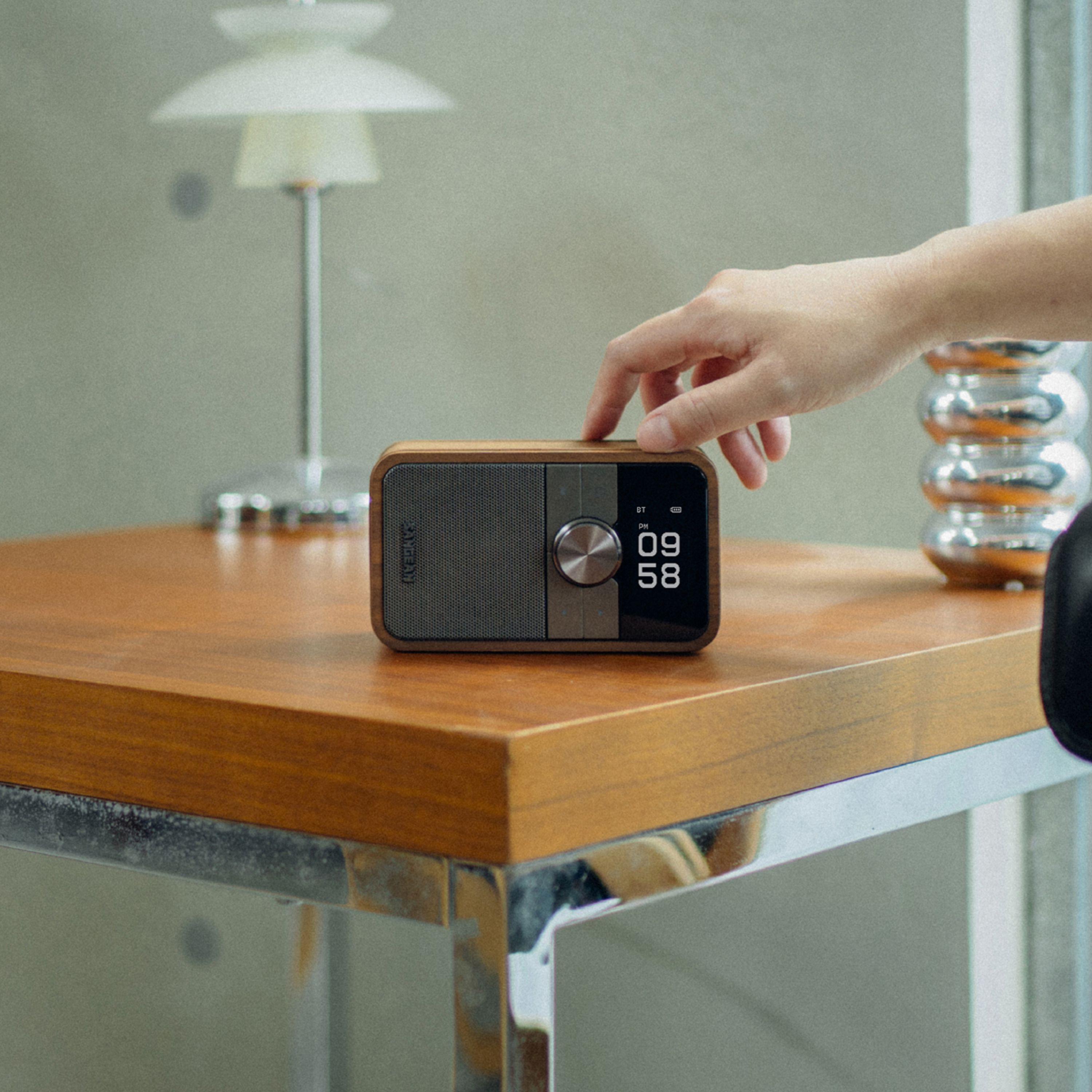 Eine Hand berührt ein Holzradio auf einem Holztisch. Eine Lampe und ein dekoratives Glasobjekt sind im Hintergrund.
