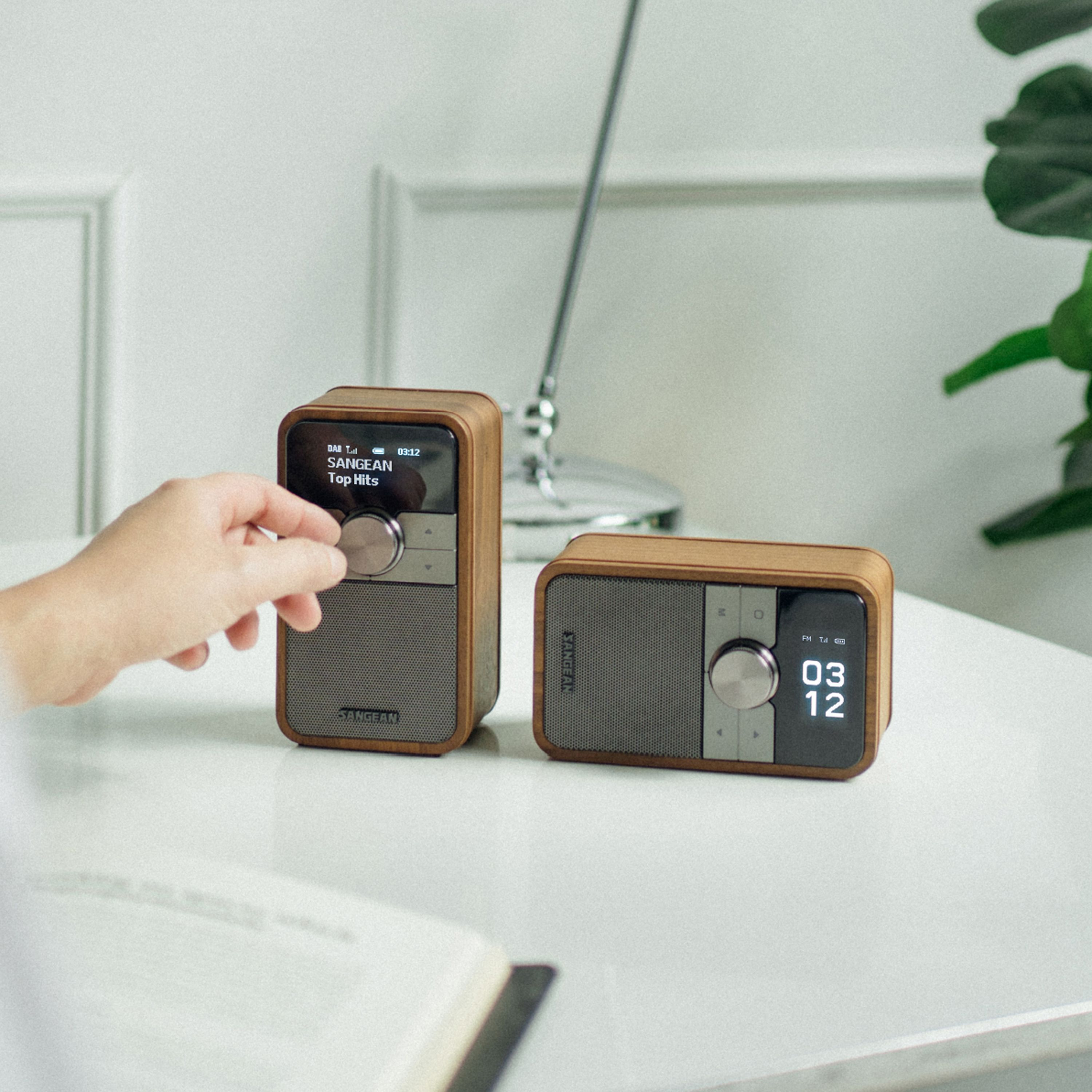 Zwei Radios mit Holzrahmen stehen auf einem Tisch. Eine Hand stellt eines ein. Der Hintergrund ist weiß.