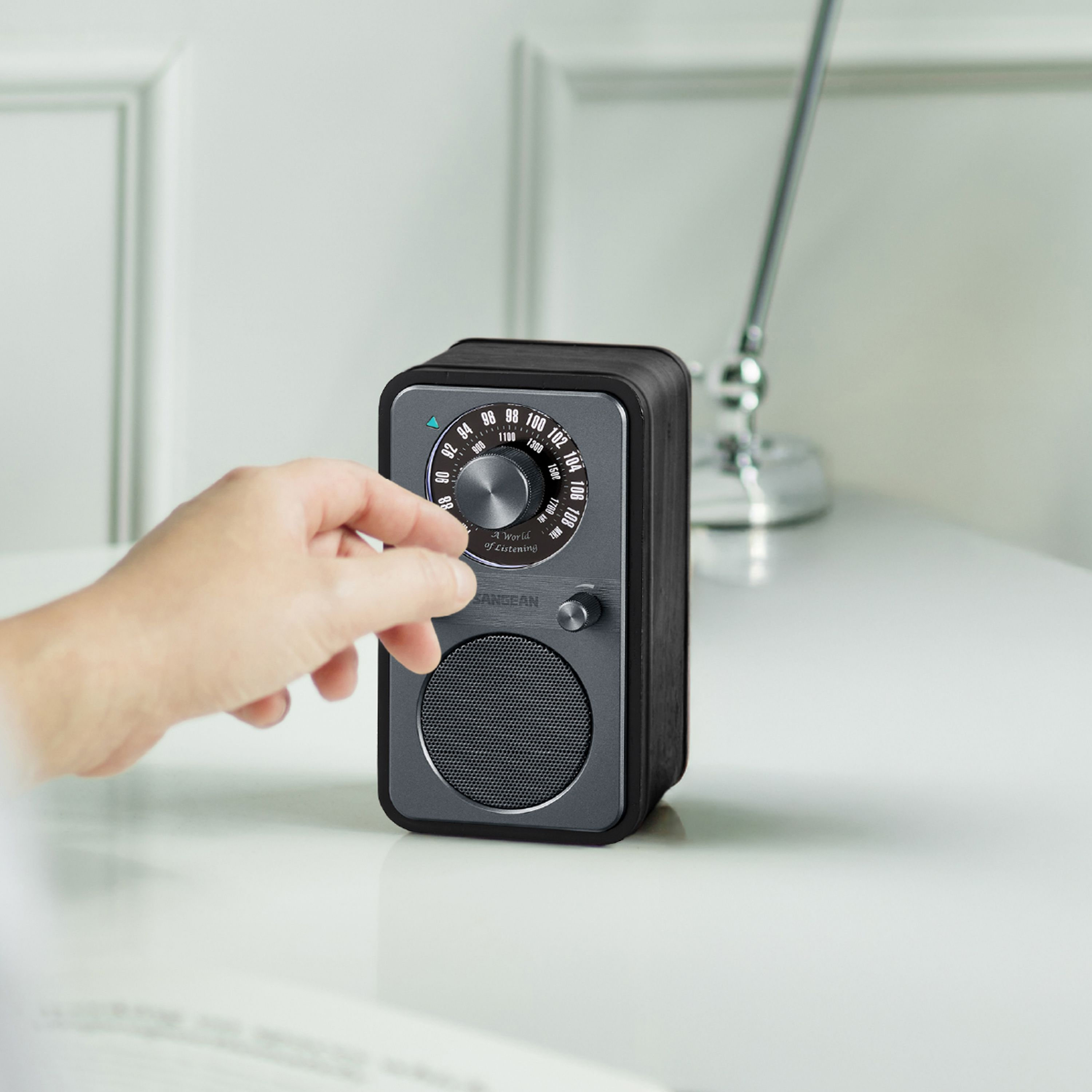 Eine Hand stellt den Drehregler eines kleinen Retro-Radios auf einer weißen Oberfläche ein. Eine metallische Antenne steht dahinter.