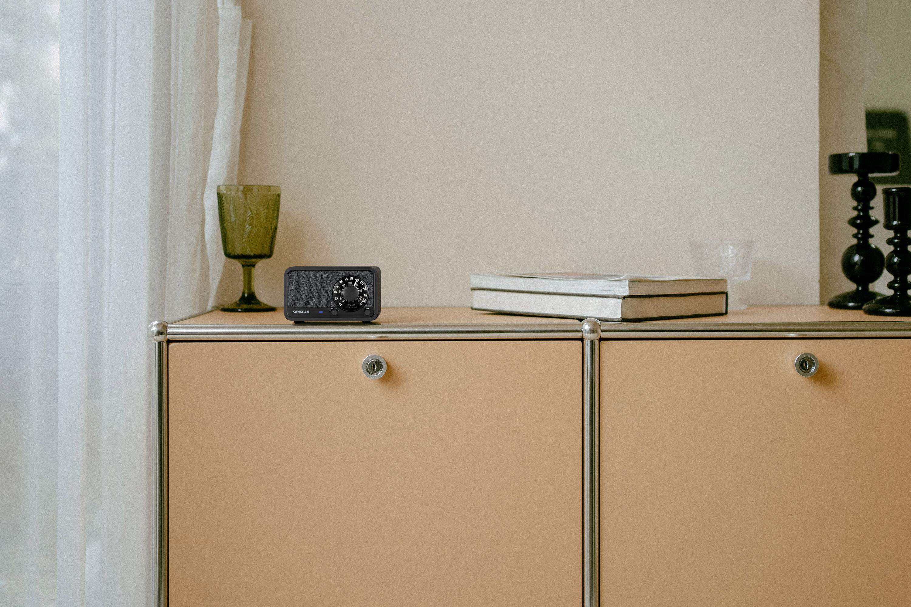 Ein beigefarbener Schrank mit einem schwarzen Radio, Büchern und Glas. Ein grünes Glas und schwarze Kerzenhalter sind ebenfalls sichtbar.