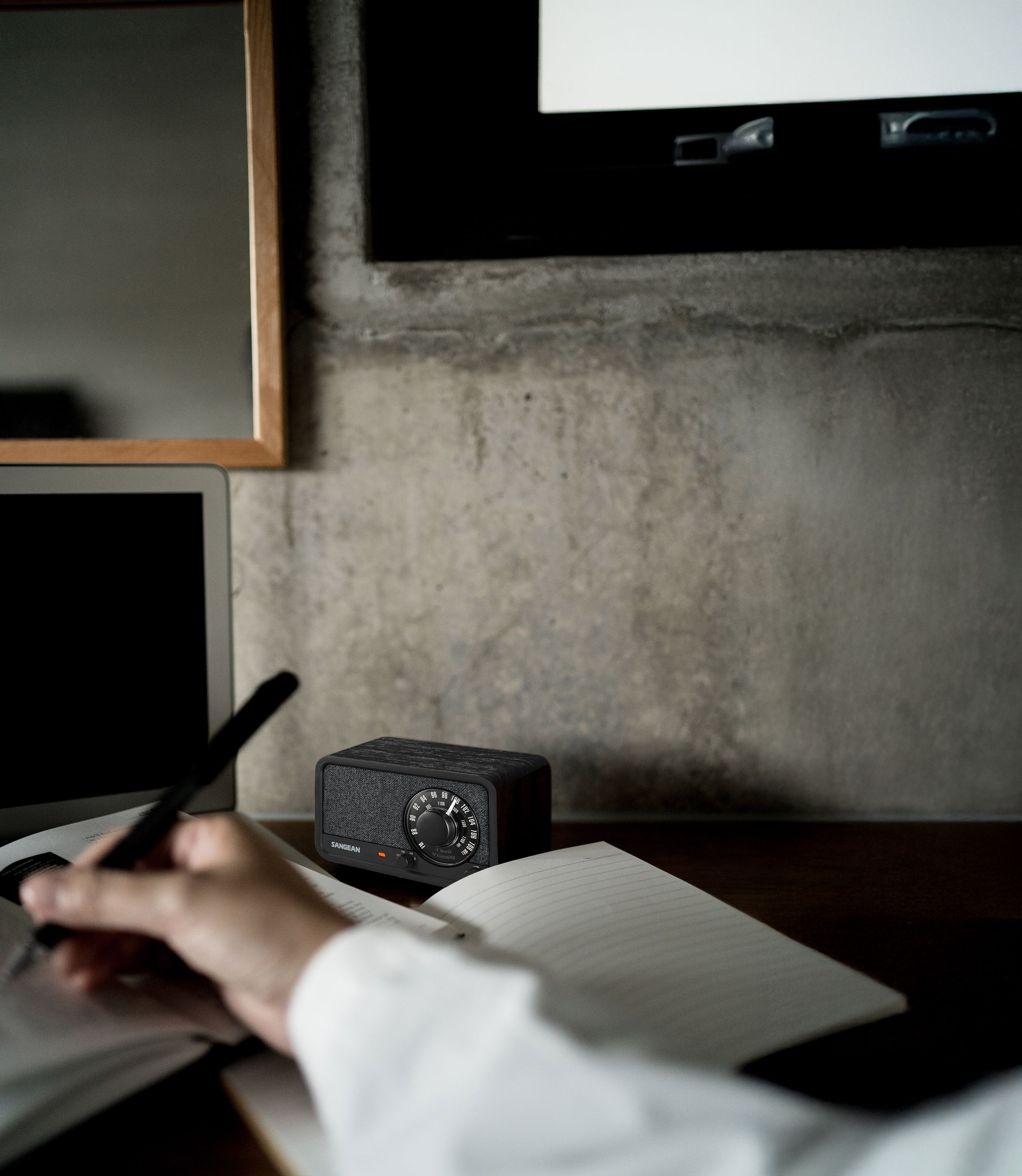 Eine Person schreibt in einem Notizbuch, ein Radio und ein Laptop stehen auf einem Holztisch. Im Hintergrund ist eine Betonmauer.
