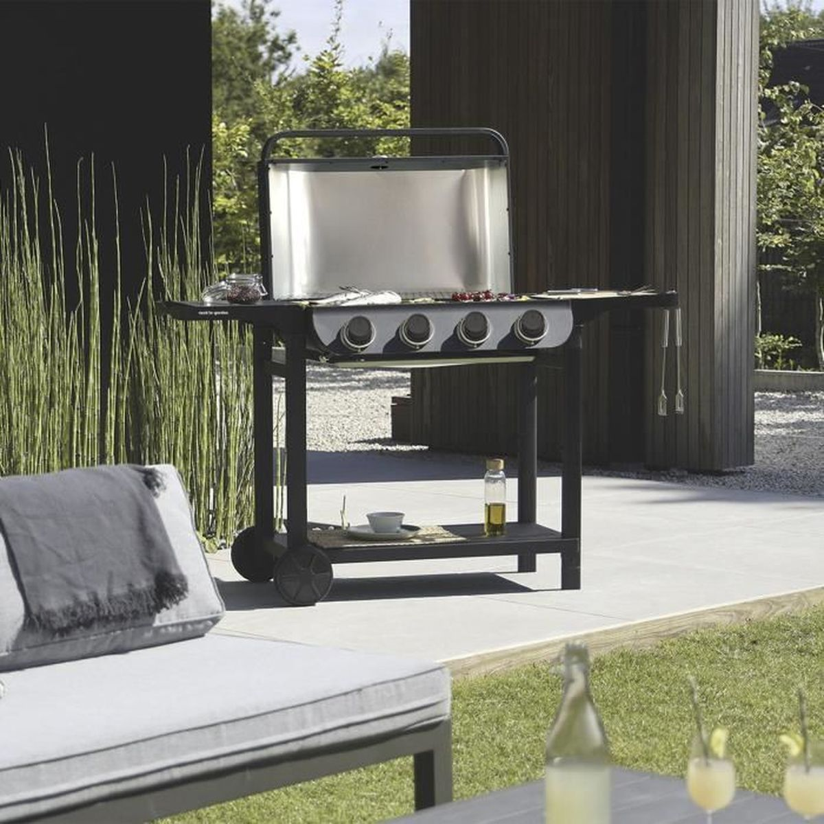 Ein moderner Außengrill auf einer Terrasse. Ein Liegestuhl steht im Vordergrund, Getränke unten rechts. Hohes Gras im Hintergrund.