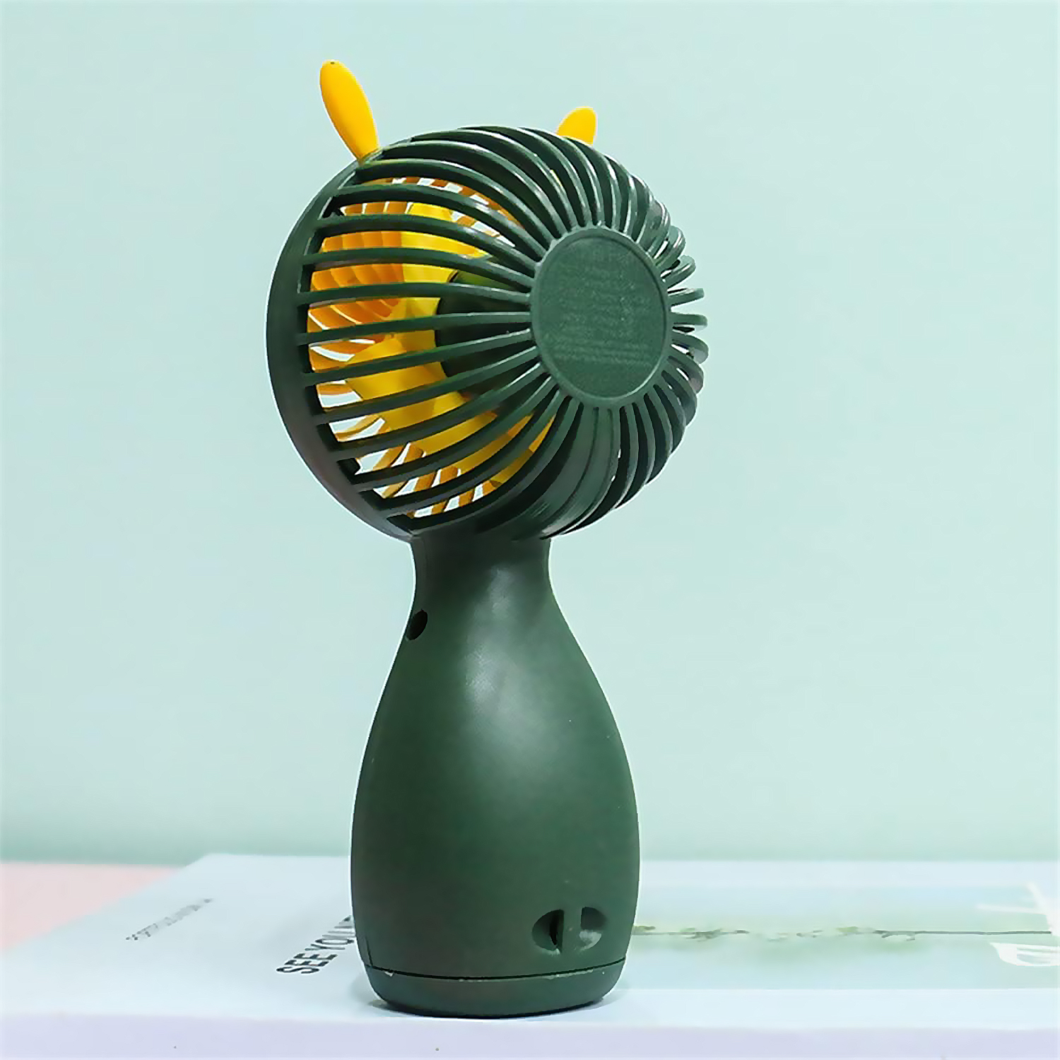 Un ventilatore portatile verde e giallo. Ha una testa rotonda con pale e un manico curvo. È su una superficie bianca.