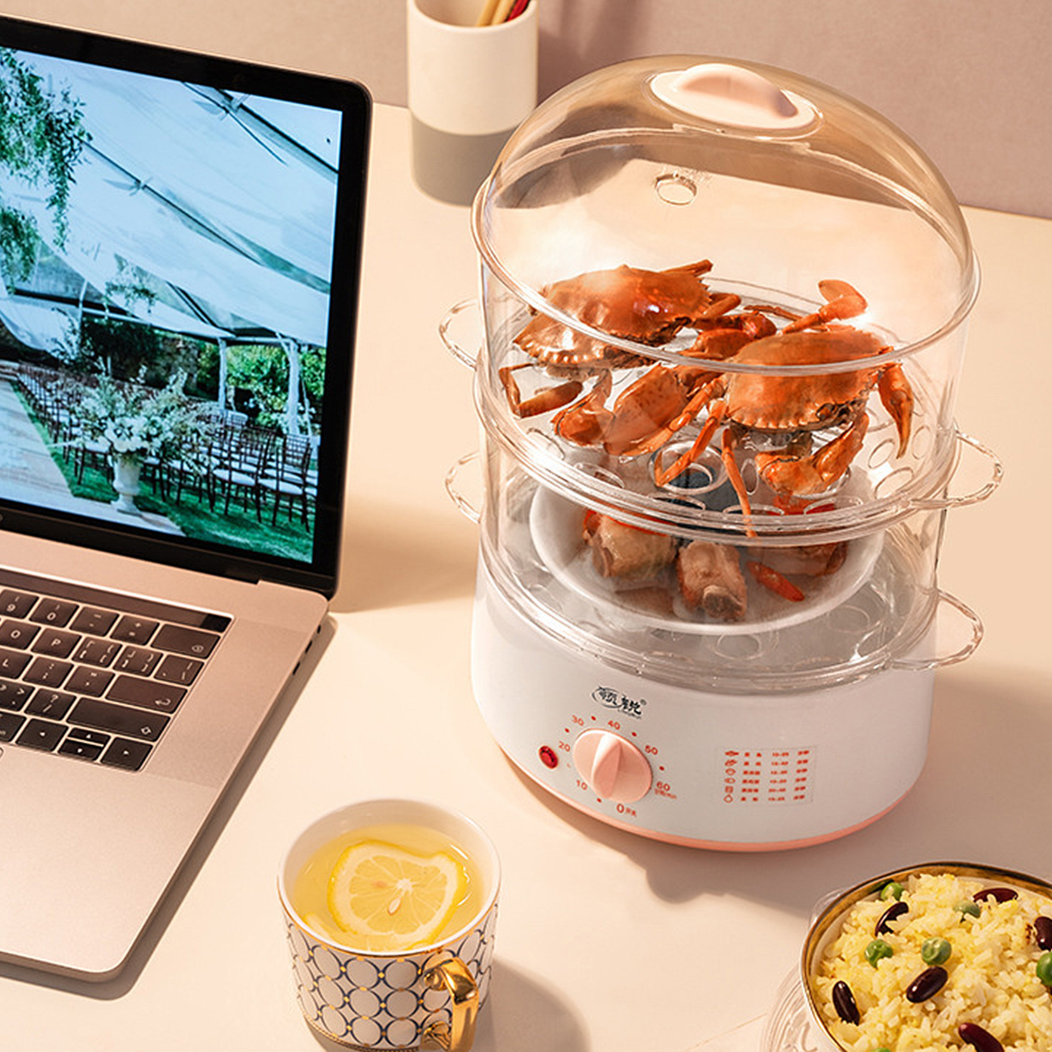 Dampfgarer mit Krabben, Laptop mit Hochzeitsszene, Tasse mit Zitrone und Reisschüssel auf einem Tisch.