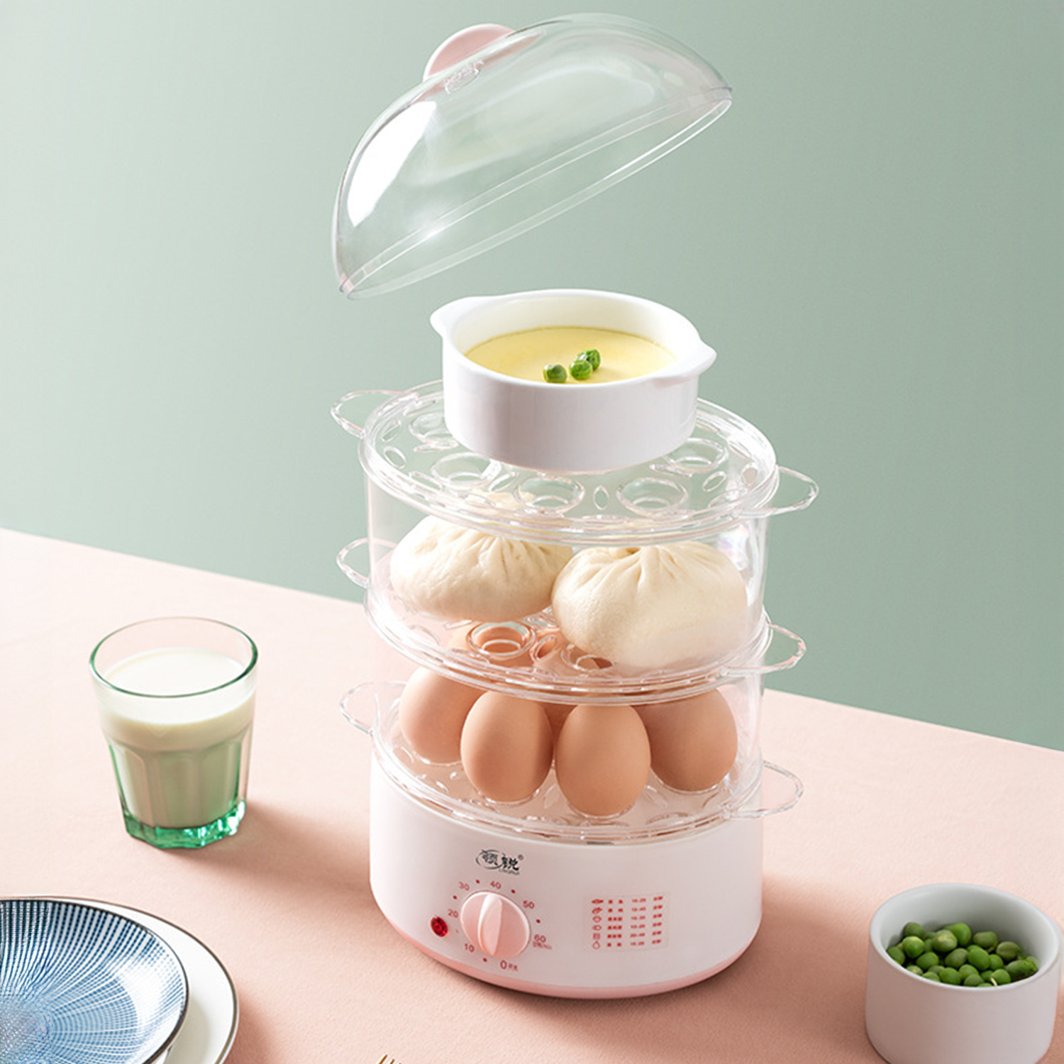 Ein Lebensmitteldämpfer mit Eiern, Brötchen und Pudding. Ein Glas Milch, eine Schüssel Erbsen und Teller stehen auf einer rosa Oberfläche.