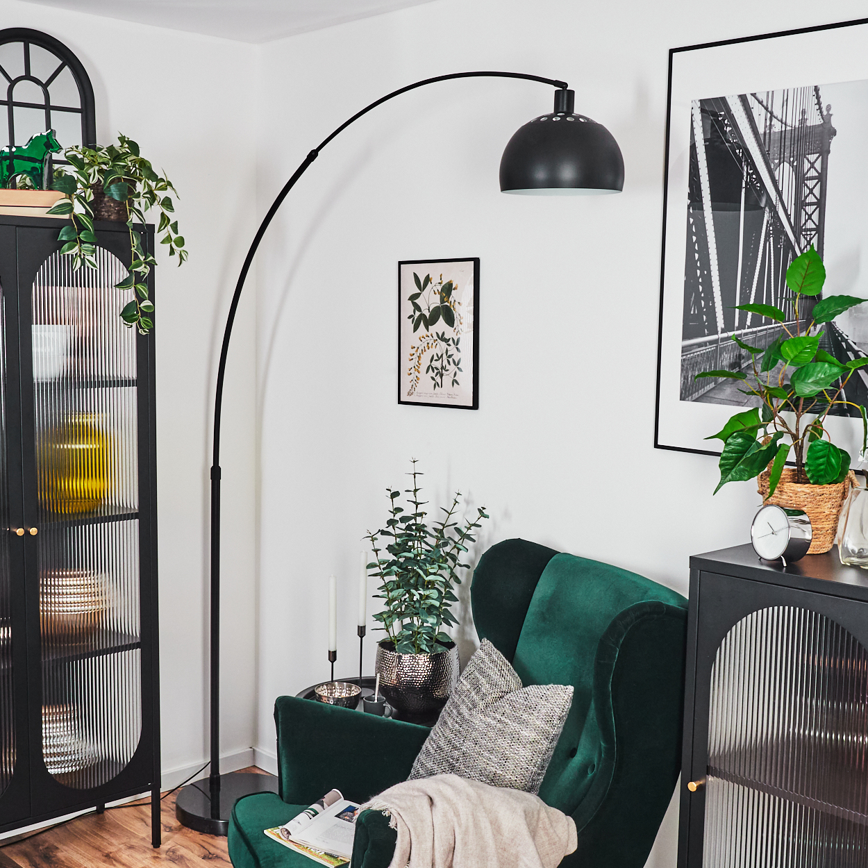 Ein Raum mit grünem Sessel, Stehlampe, schwarzem Schrank und gerahmter Kunst. Pflanzen sind überall platziert. Die Szene ist gut beleuchtet.