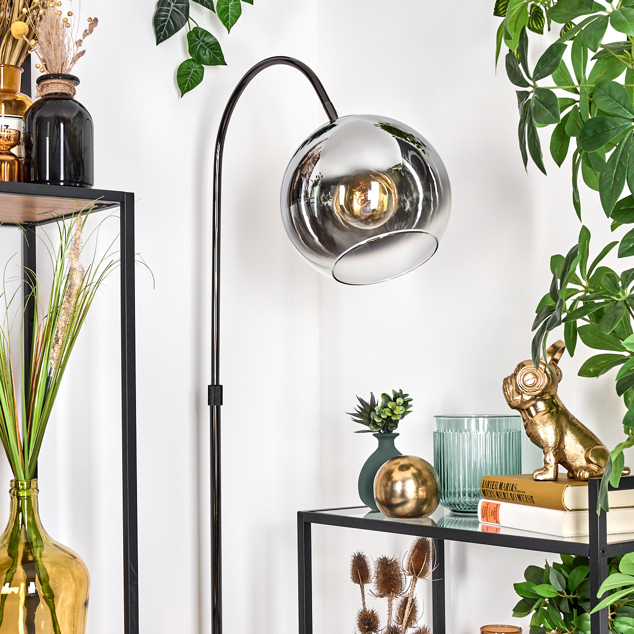 Eine moderne Lampe mit silbernem Kugelschirm. Regale beherbergen dekorative Gegenstände, darunter Vasen, eine goldene Bulldoggenstatue und Bücher.