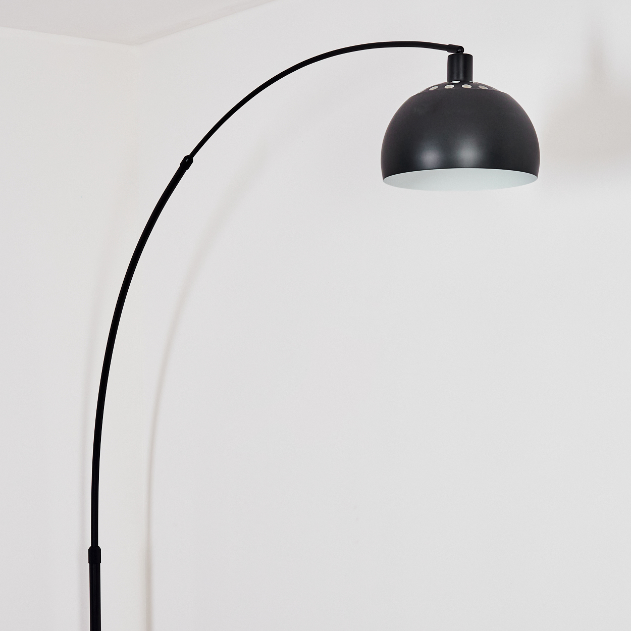 Eine schwarze Bogenlampe steht vor einer weißen Wand. Die Lampe hat einen kuppelförmigen Kopf mit weißem Innenraum.