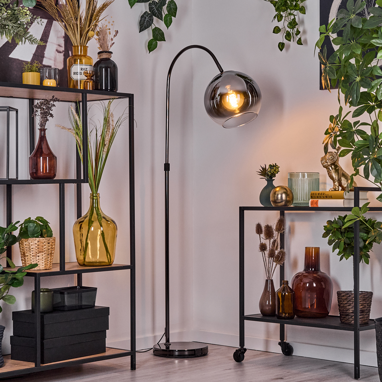 Wohnzimmer mit Regalen, Pflanzen und einer schwarzen Stehlampe mit rundem Lampenschirm. Verschiedene Vasen und Dekorationen sind vorhanden.