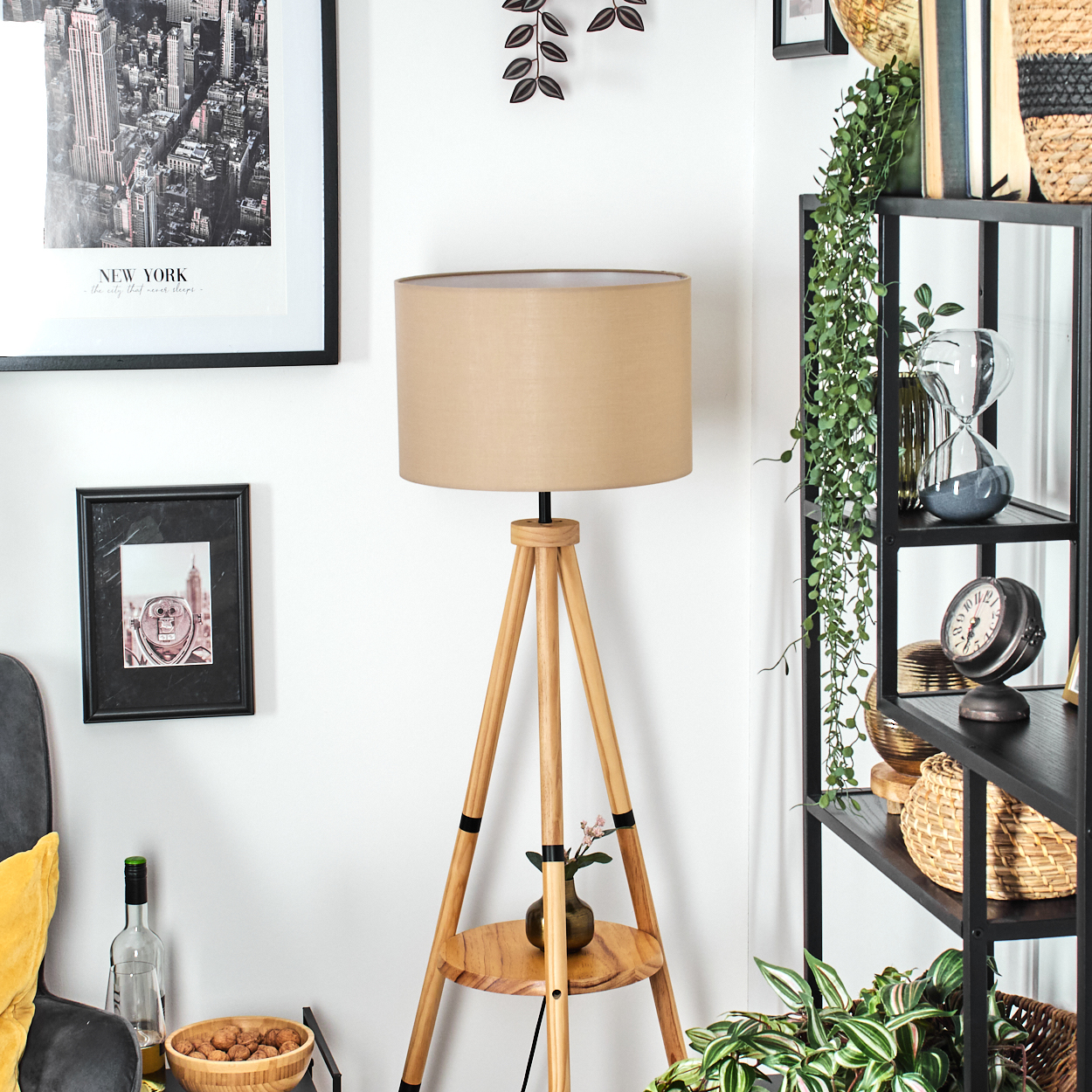 Ein Zimmer mit einer Holzlampe, schwarzen Regalen und Bildern. Die Lampe hat einen beigen Schirm. Es gibt Pflanzen, eine Uhr und eine Schüssel mit Nüssen.