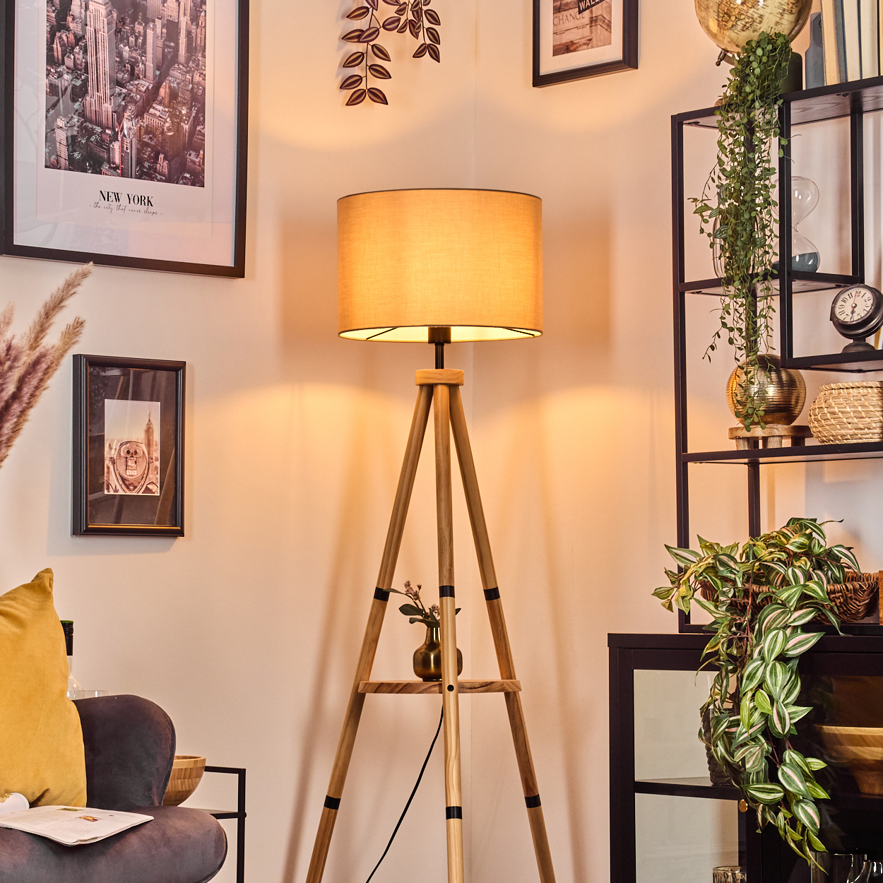 Ein Raum mit einer hölzernen Stativlampe, gerahmten Bildern, einem schwarzen Regal mit Pflanzen und einem Sofa mit gelbem Kissen.