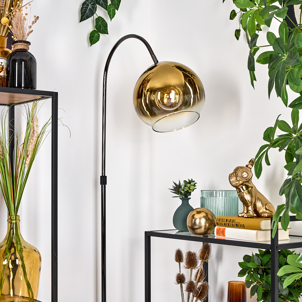 Eine goldene Lampe mit Pflanzen im Hintergrund. Ein Regal enthält Dekorationen wie eine Bulldoggen-Statue und Bücher.