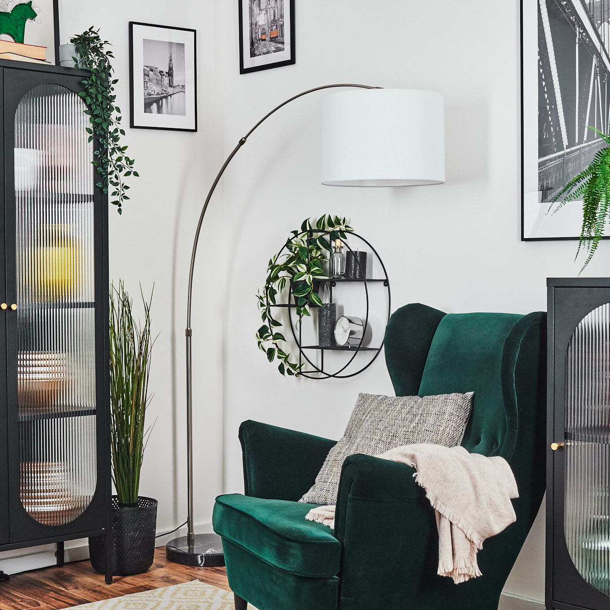 Ein grüner Sessel mit Decke und Kissen, ein schwarzes Regal mit Deko und eine hohe Lampe stehen in einem Raum mit Kunst.
