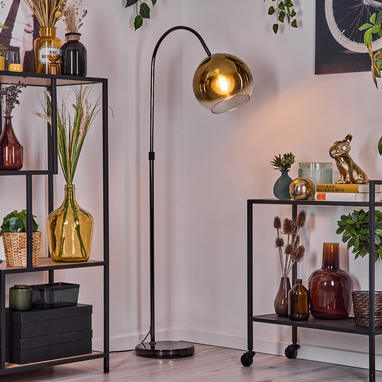 Raumecke mit schwarzem Metallregal und Stehlampe. Vasen mit Pflanzen und Dekorationen stehen auf den Regalen. Eine goldene Lampe leuchtet.