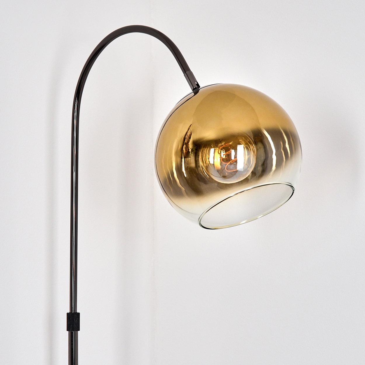 Eine moderne Stehlampe mit einem gebogenen schwarzen Metallarm und einem runden, gold getönten Glasschirm. Die Glühbirne ist sichtbar. Der Hintergrund ist weiß.