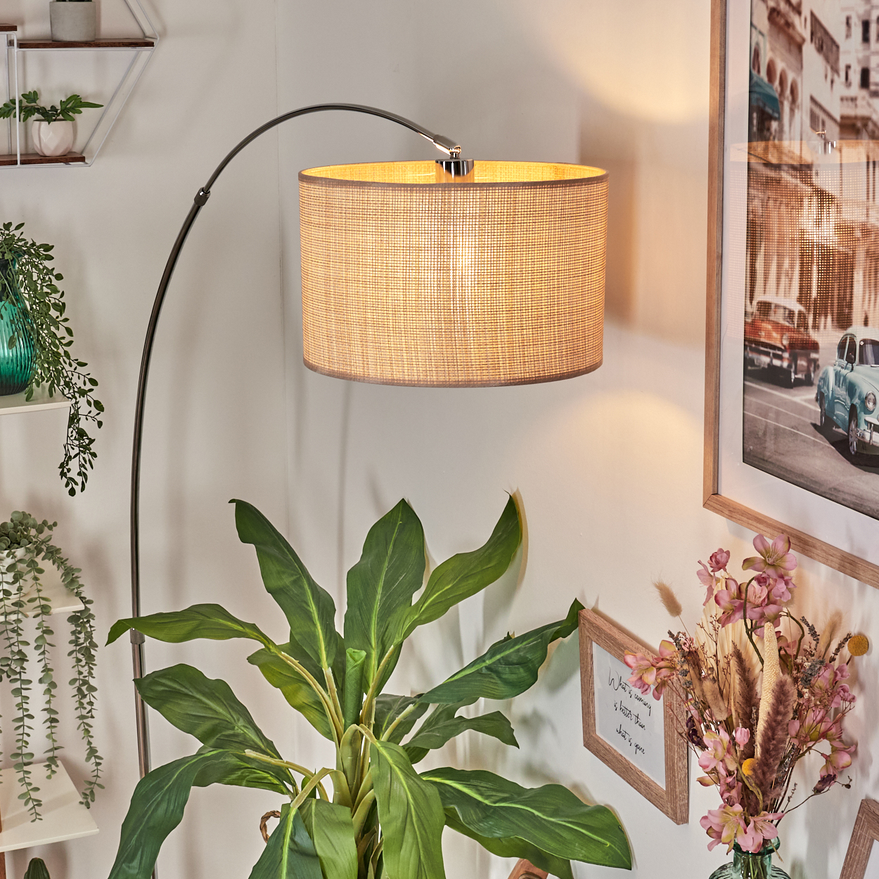 Eine Stehlampe mit gewebtem Schirm, eine große grüne Pflanze, ein gerahmtes Bild und ein kleines Schild mit Blumen.