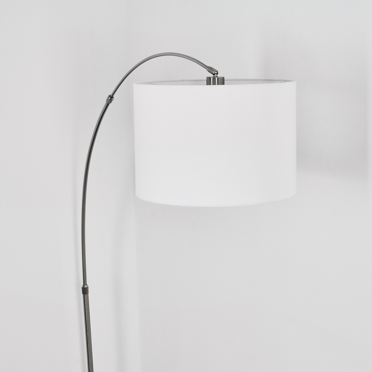 Eine moderne Bogen-Stehlampe mit weißem Lampenschirm. Der Metallarm der Lampe ist gebogen, vor einer weißen Wand.