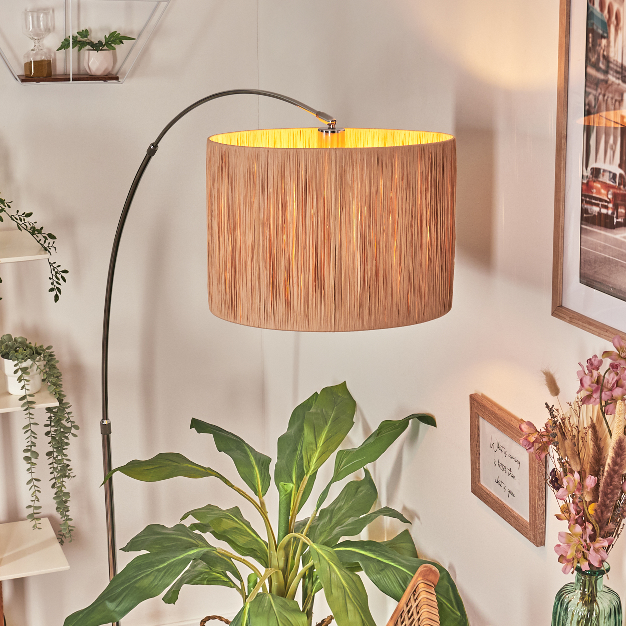 Eine Stehlampe mit beigem Lampenschirm hängt über einer Pflanze, mit einem Bilderrahmen und einer Blumenvase im Hintergrund.