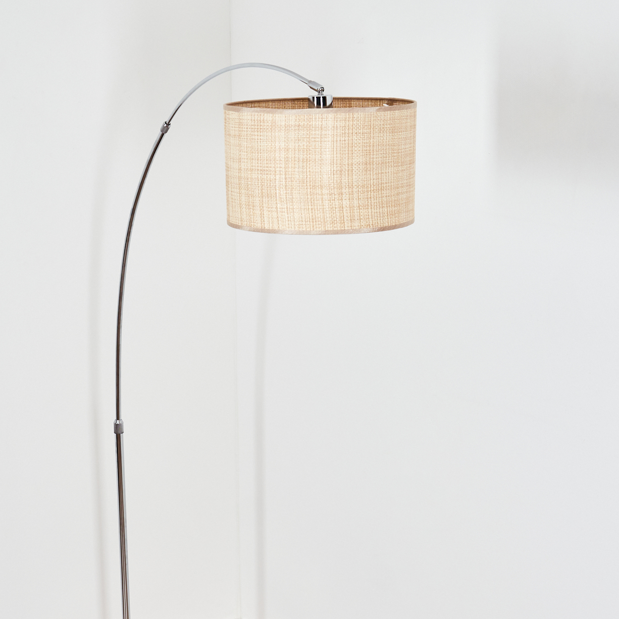 Eine moderne Bogen-Stehlampe mit beigem Lampenschirm steht vor einer weißen Wand. Die Lampe hat ein Metallgestell.