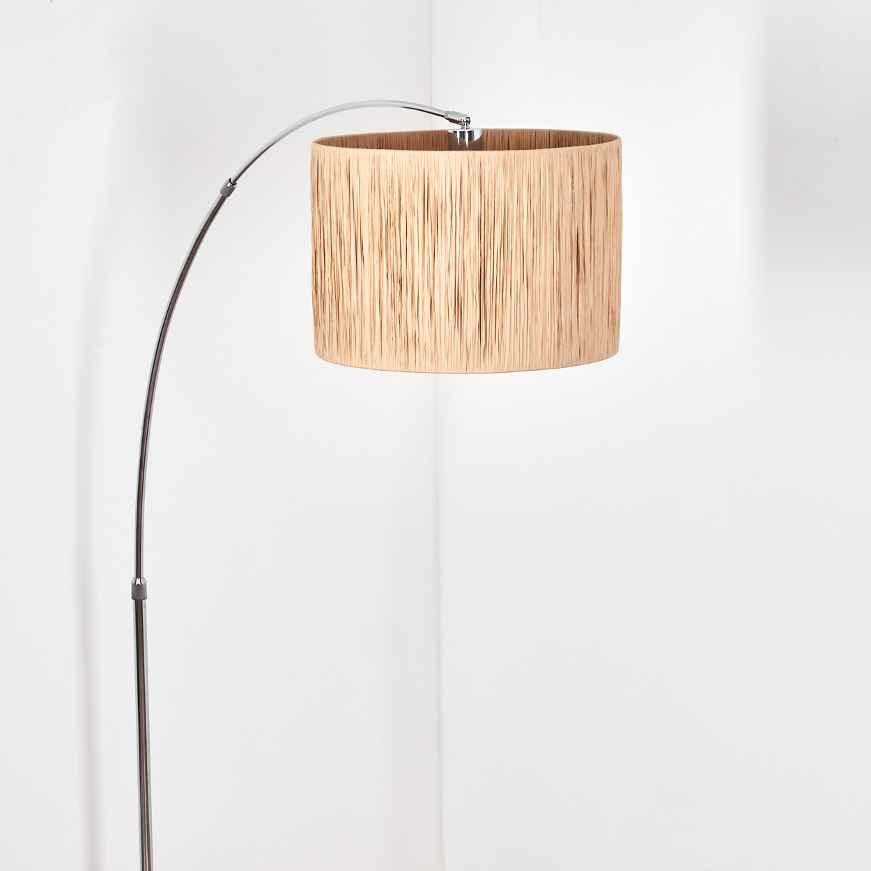 Moderne Bogen-Stehlampe. Die Lampe hat einen beigefarbenen Schirm und ein silbernes Metallgestell, vor einer weißen Wand.