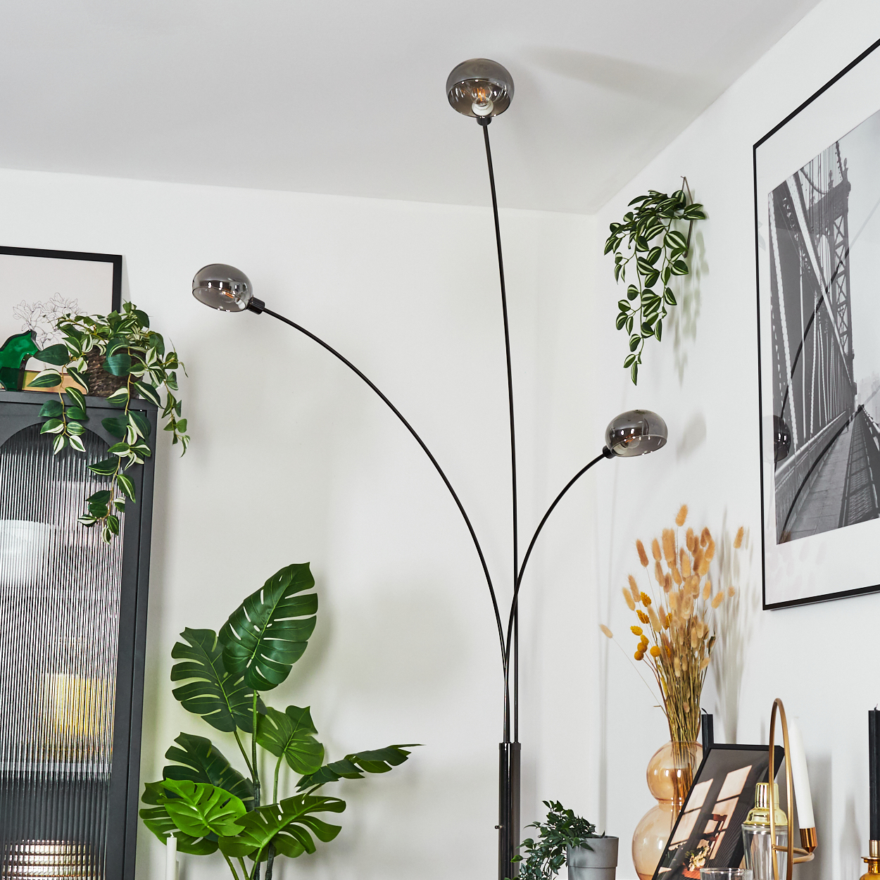 Eine moderne Stehlampe mit drei Glaskugellichtern. Pflanzen und ein gerahmtes Brückenkunstwerk im Hintergrund.